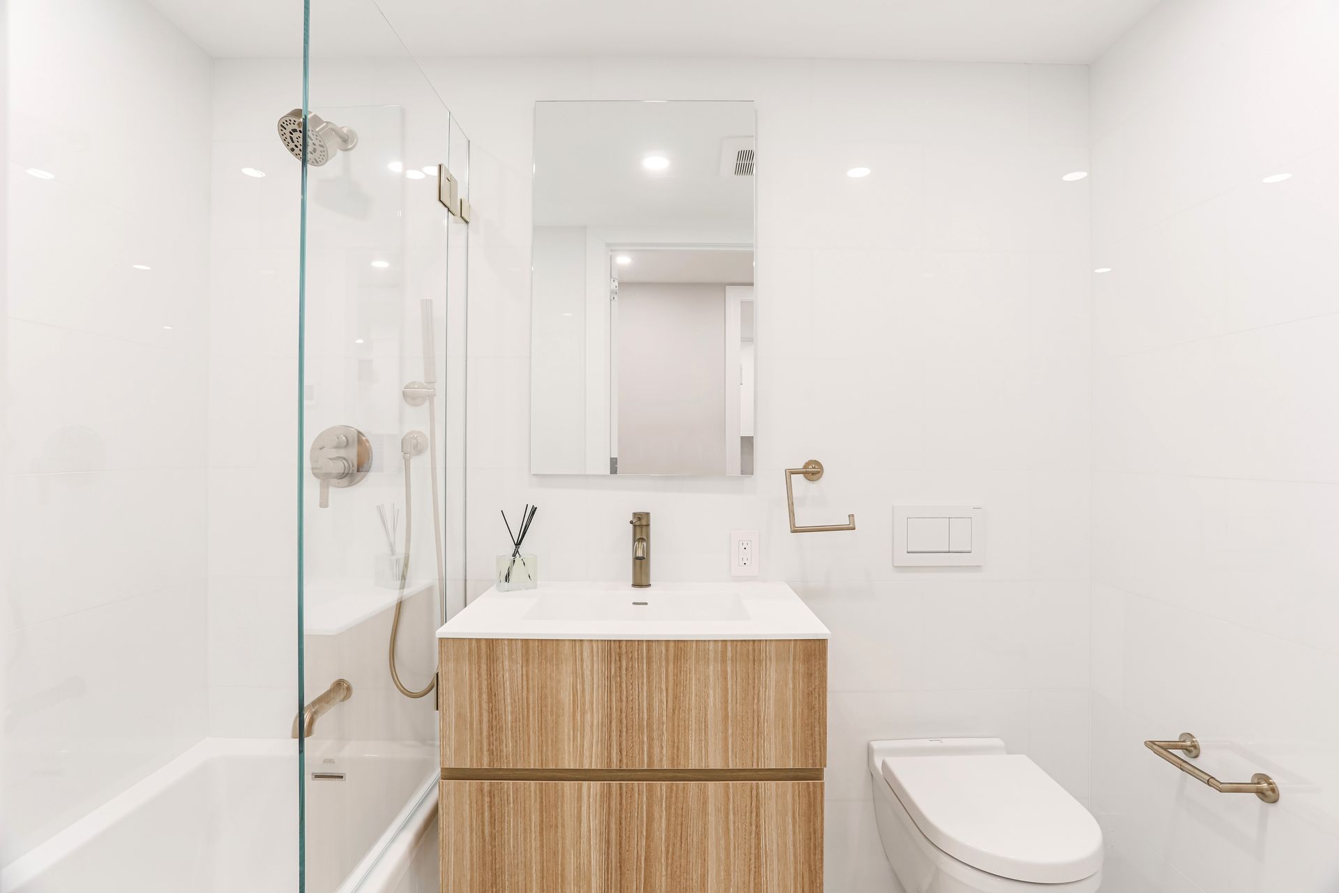 A clean, modern bathroom with a wood-toned vanity, white walls, a walk-in shower with glass doors, and a white toilet.