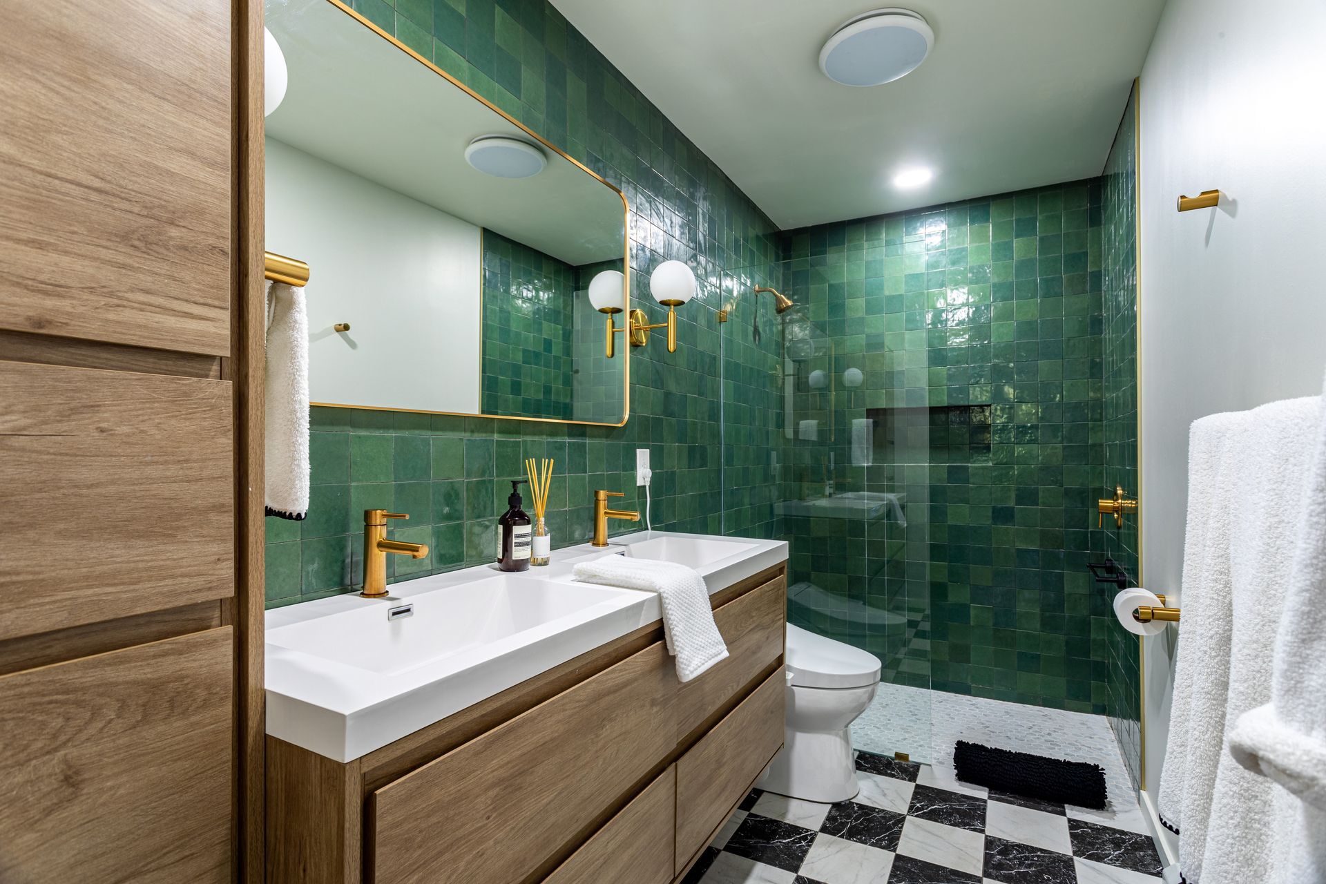 Modern bathroom with wood vanity, white countertop, gold faucets, and green tiled walls in the shower and vanity area.