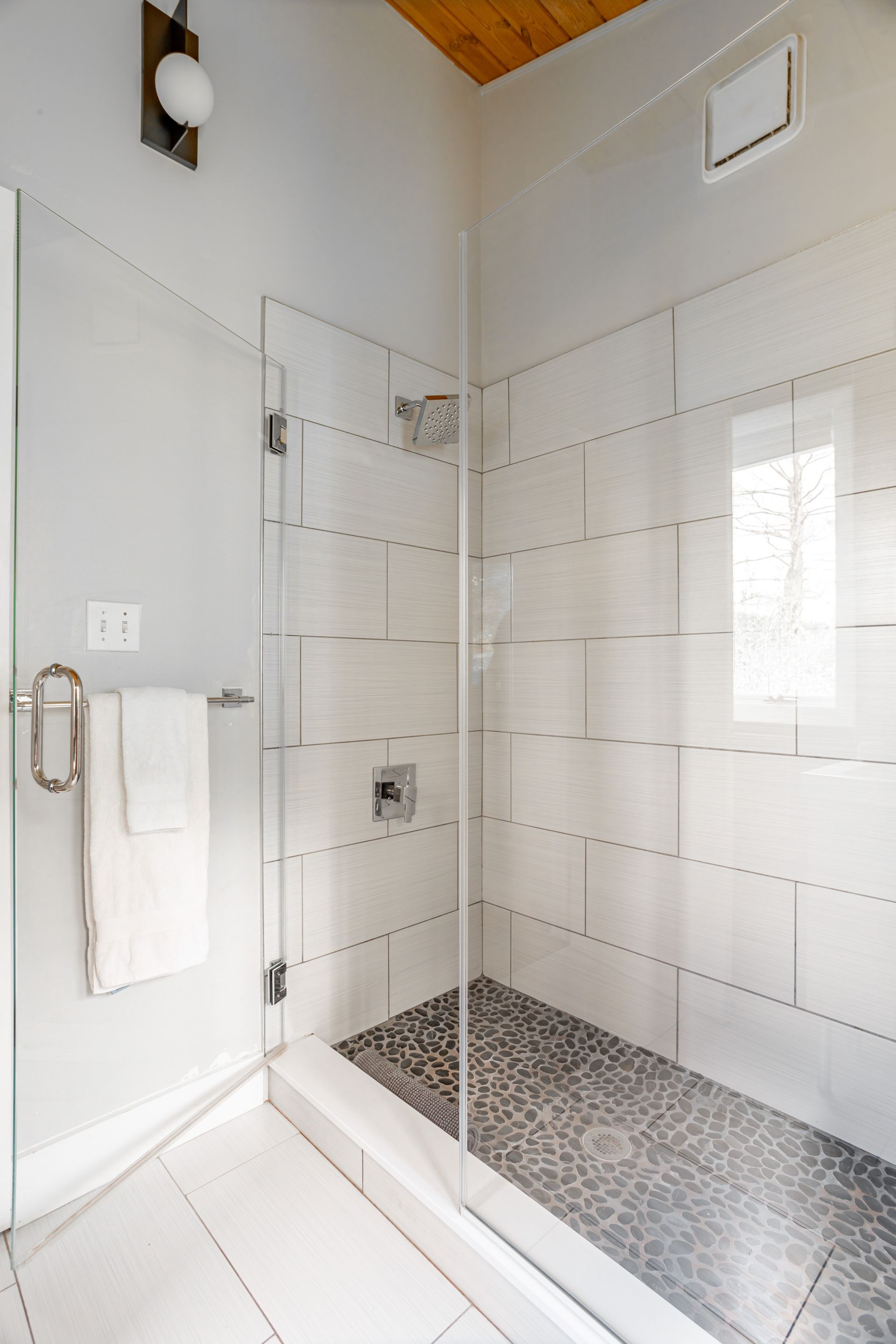 A tiled walk-in shower with a glass door, pebble floor, and a white towel hanging on a bar outside.