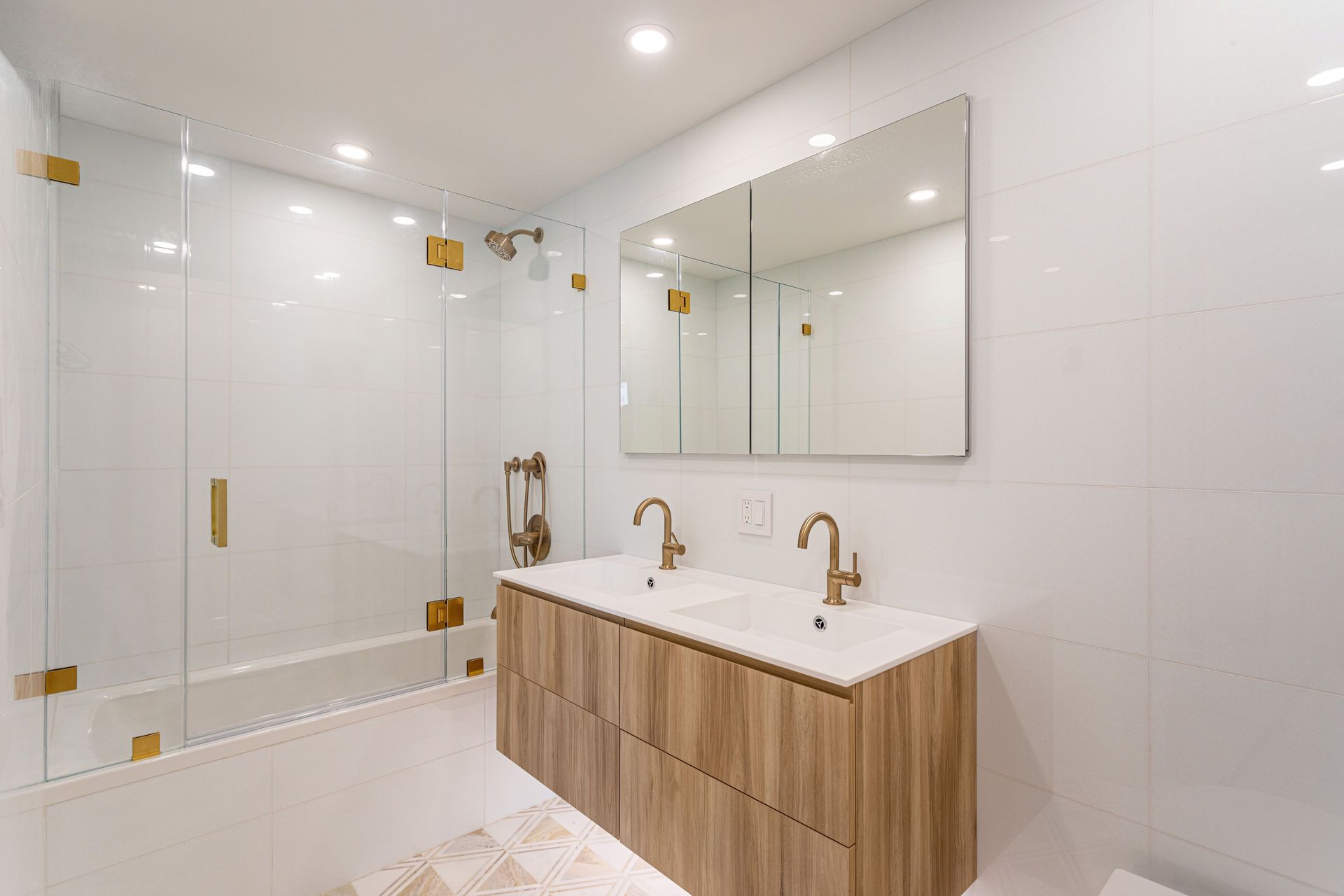 A modern bathroom with a glass-enclosed shower, a double vanity with wood-grain cabinets, and gold fixtures.