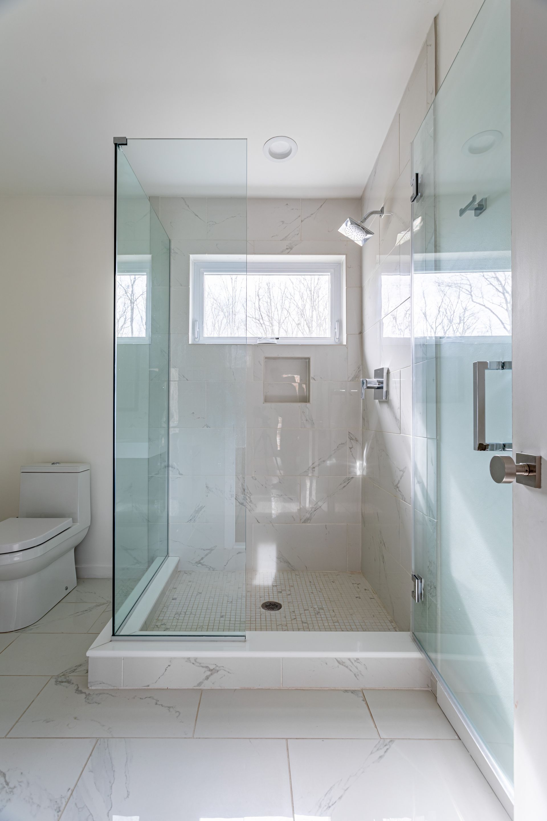 A modern walk-in shower with white marble-style tile, a glass door, a wall-mounted showerhead, and a small recessed niche.