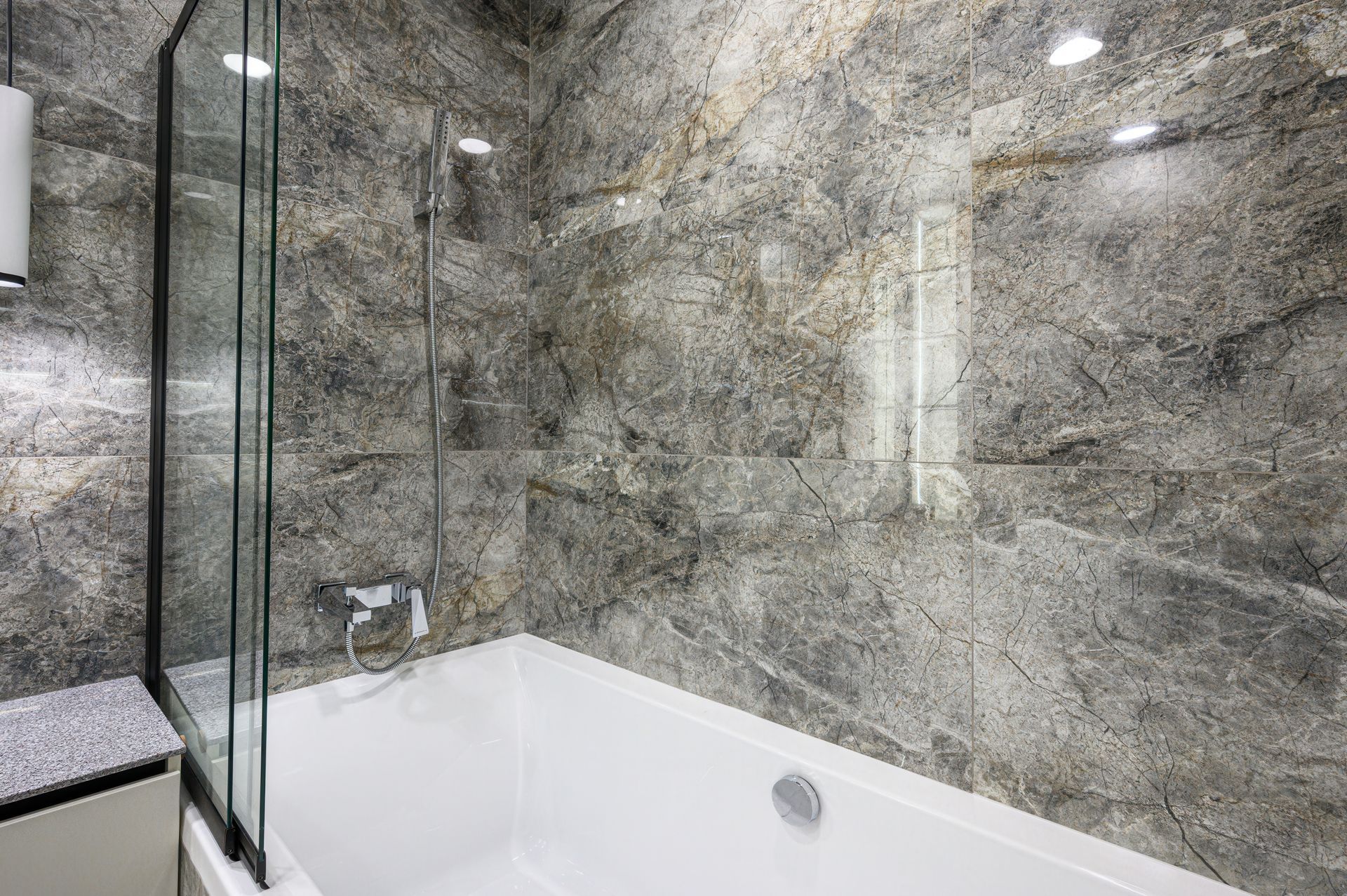 A bathtub with a glass shower screen against a wall of grey, polished stone tiles and a wall-mounted shower fixture.