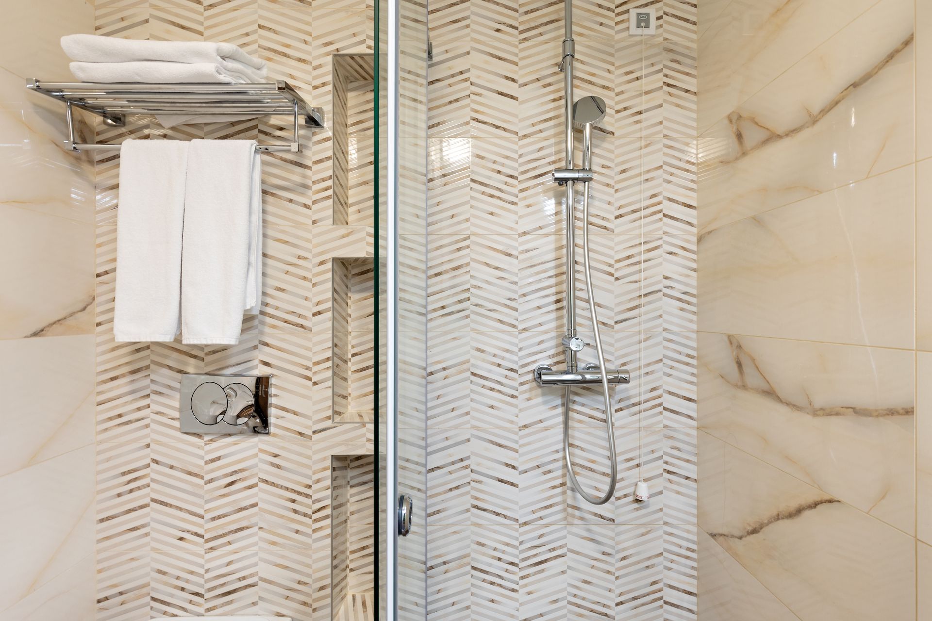 A modern shower with beige chevron-patterned wall tiles, a silver shower head, recessed shelves, and white towels.