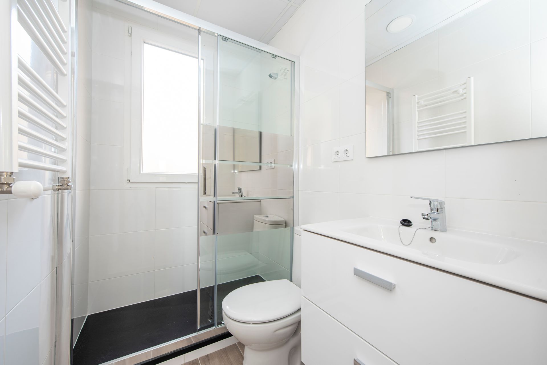A bright, modern white bathroom featuring a glass shower stall, a white vanity with a sink, and a toilet.