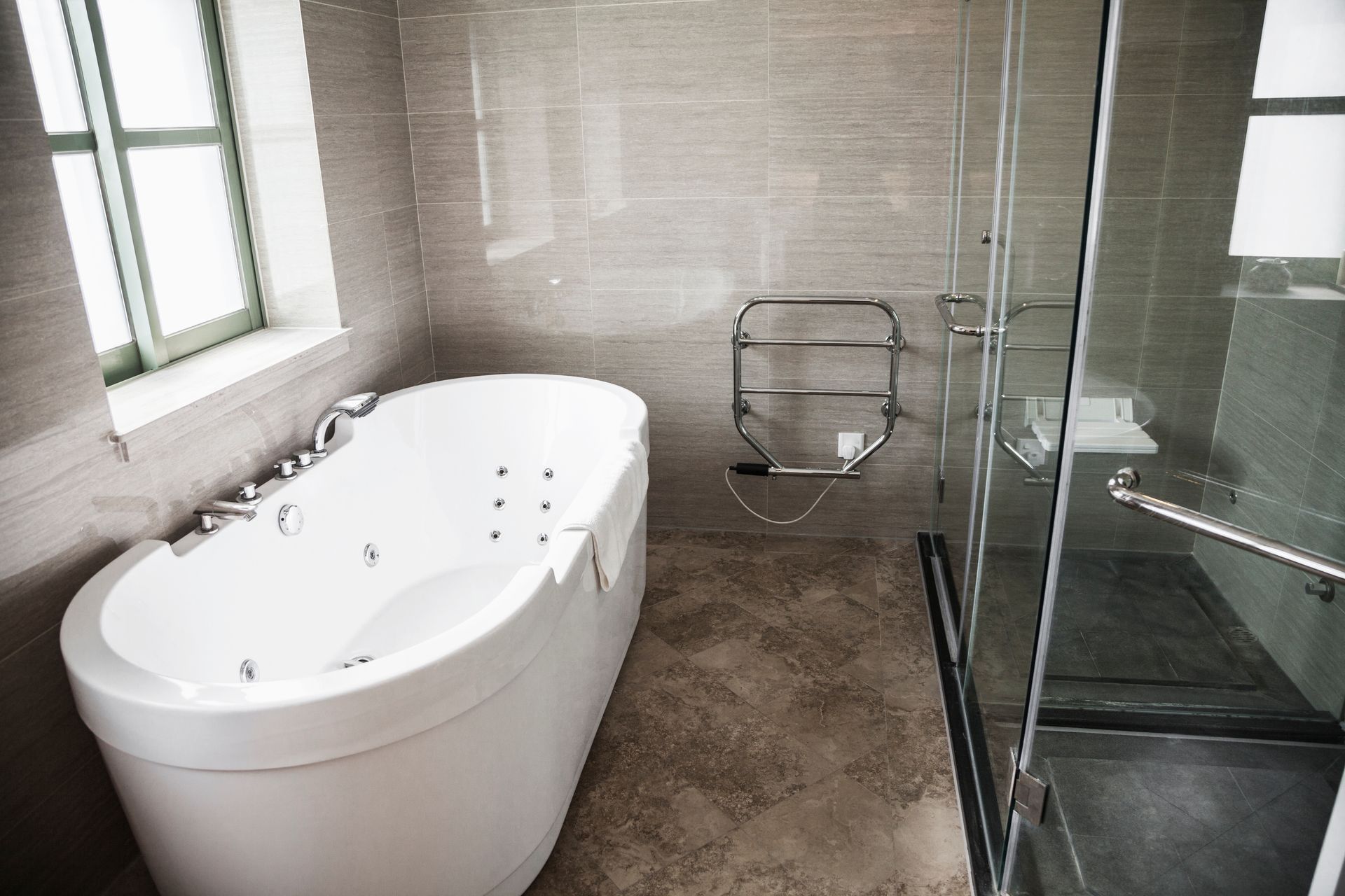 A modern bathroom with a white oval jetted tub next to a glass-enclosed shower, set against grey tiled walls and floor.