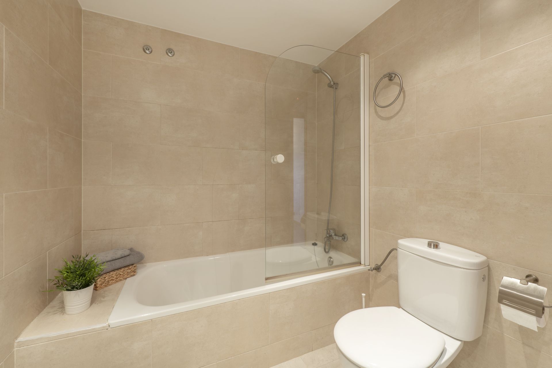 A modern bathroom featuring a white bathtub with a glass shower screen, light beige tiled walls, and a white toilet.