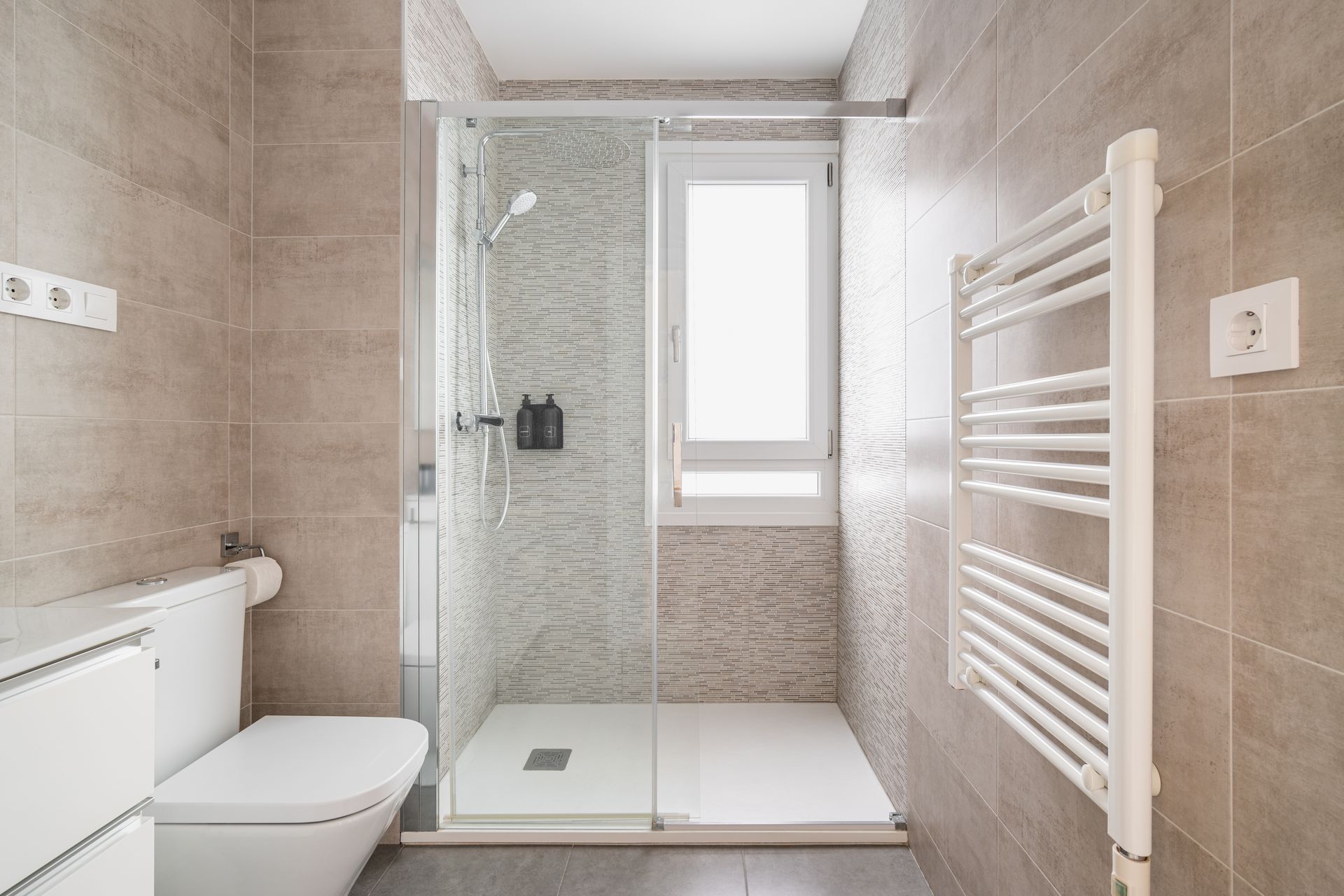 A modern bathroom with a glass-enclosed shower, a wall-mounted white toilet, and a white towel warmer on a tiled wall.
