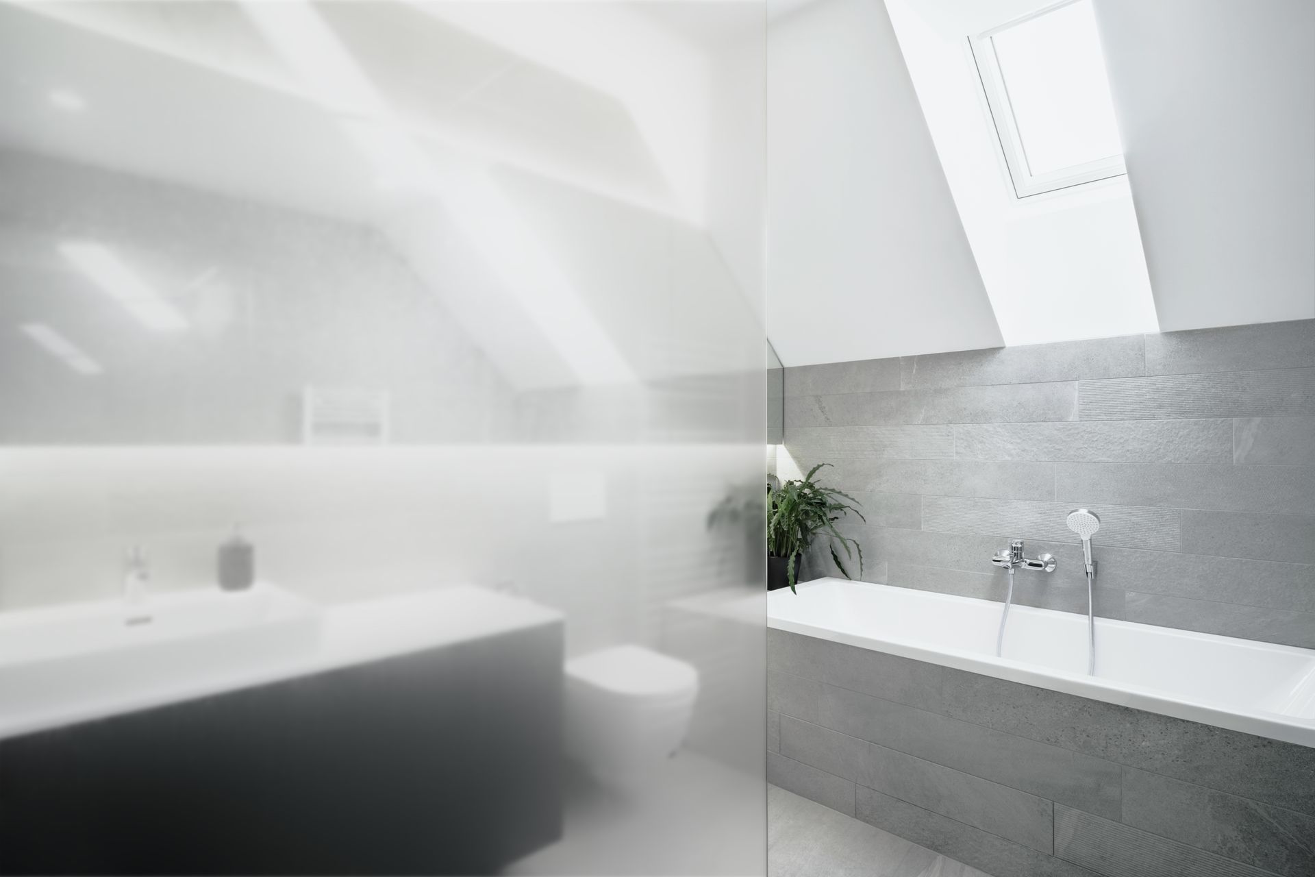 A modern, bright bathroom with a white bathtub against a grey stone wall, a sink area, and a large frosted glass partition.