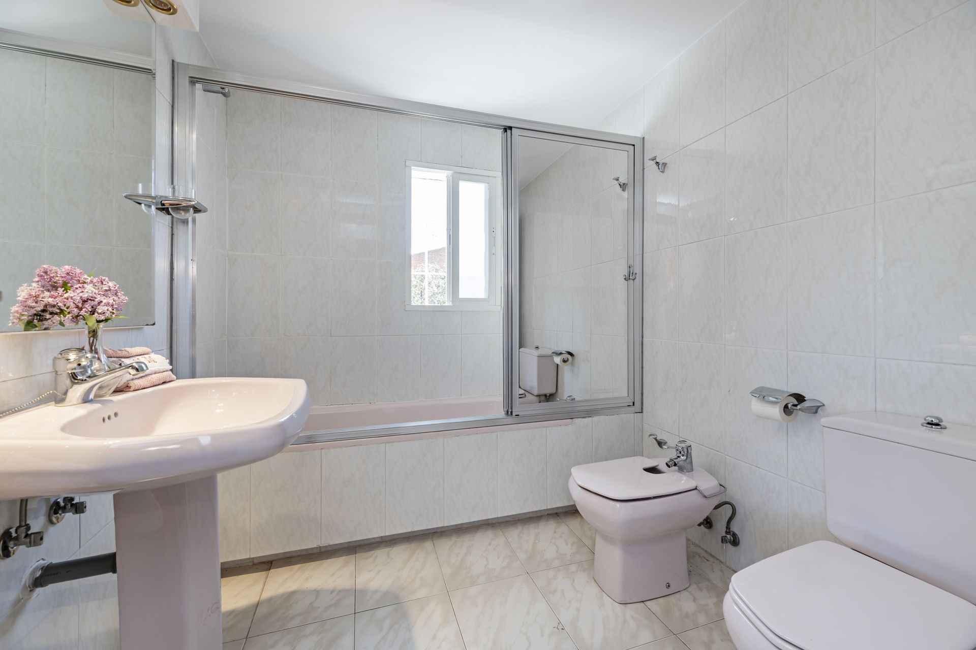 A brightly lit bathroom with white tiled walls, a pedestal sink, a bidet, a toilet, and a glass-enclosed bathtub.