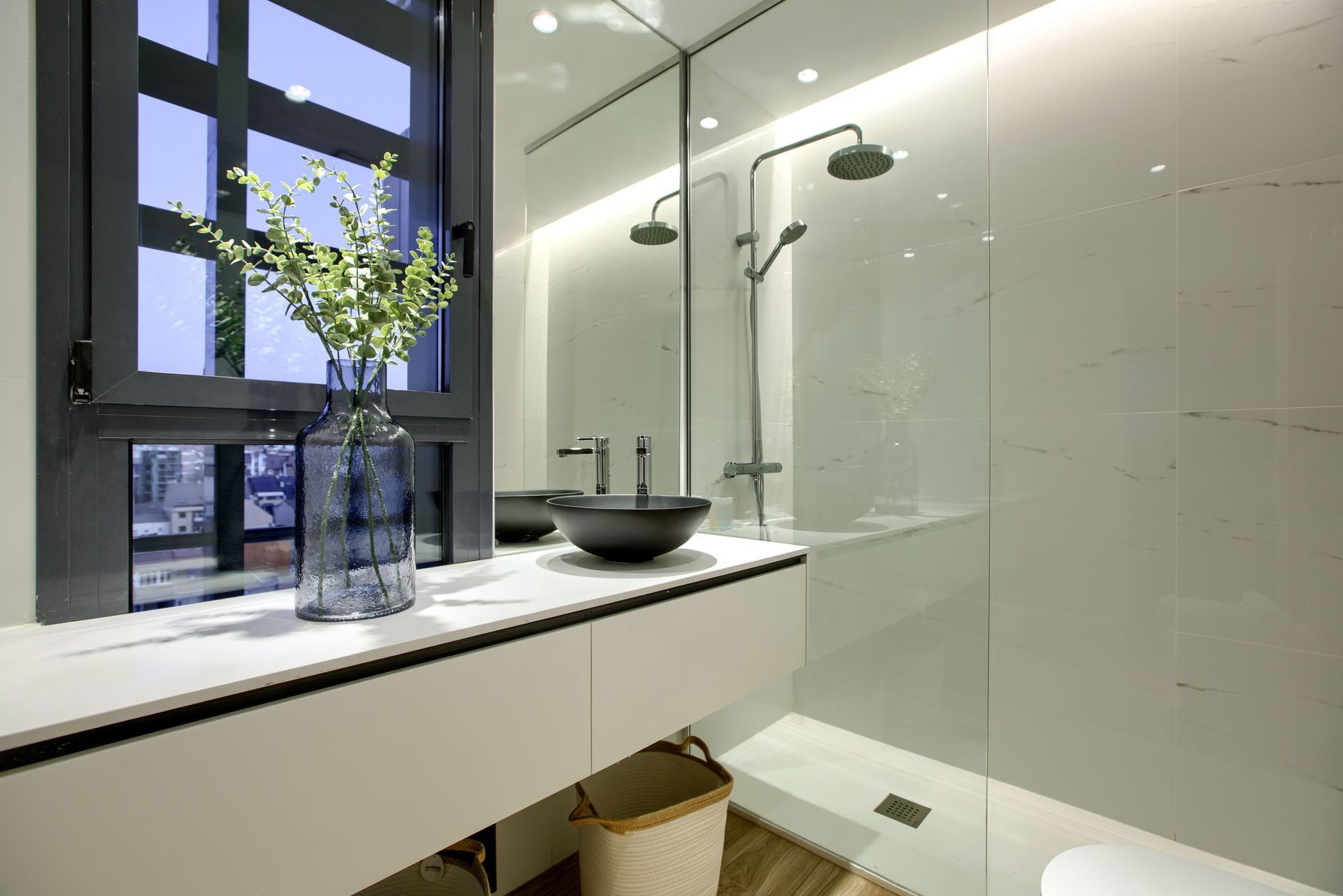 A modern bathroom featuring a white floating vanity, a black vessel sink, a tall clear vase with greenery, and a shower.