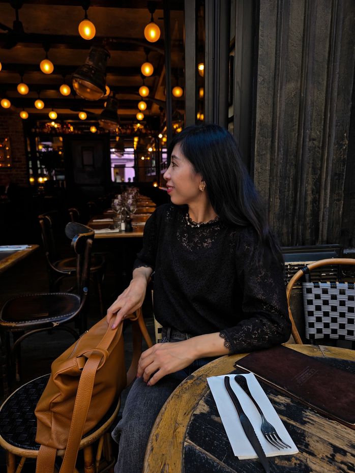 一位女士坐在咖啡桌旁，目光看向一旁。她身穿黑色上衣，手提一個棕色包包。室內光線昏暗，點綴著串燈。