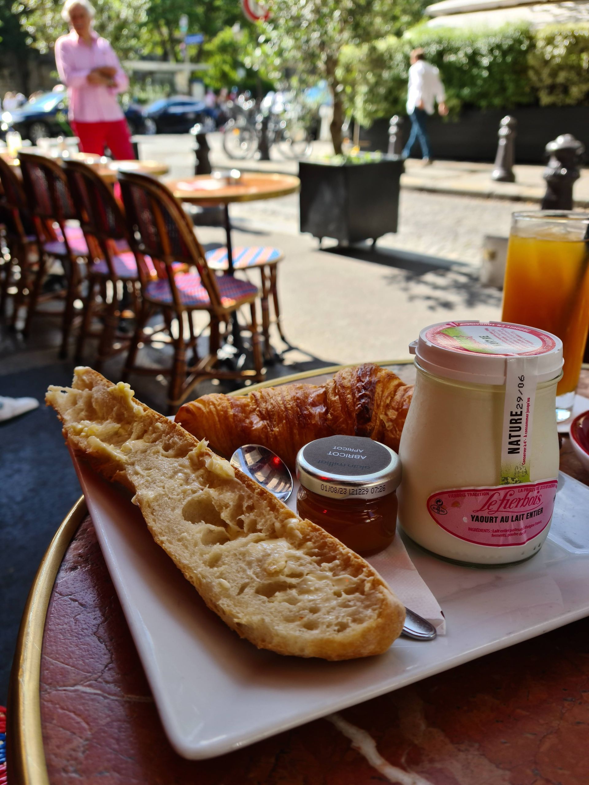 早餐拼盤：麵包、可頌、優格、果醬、柳橙汁。戶外咖啡館環境。