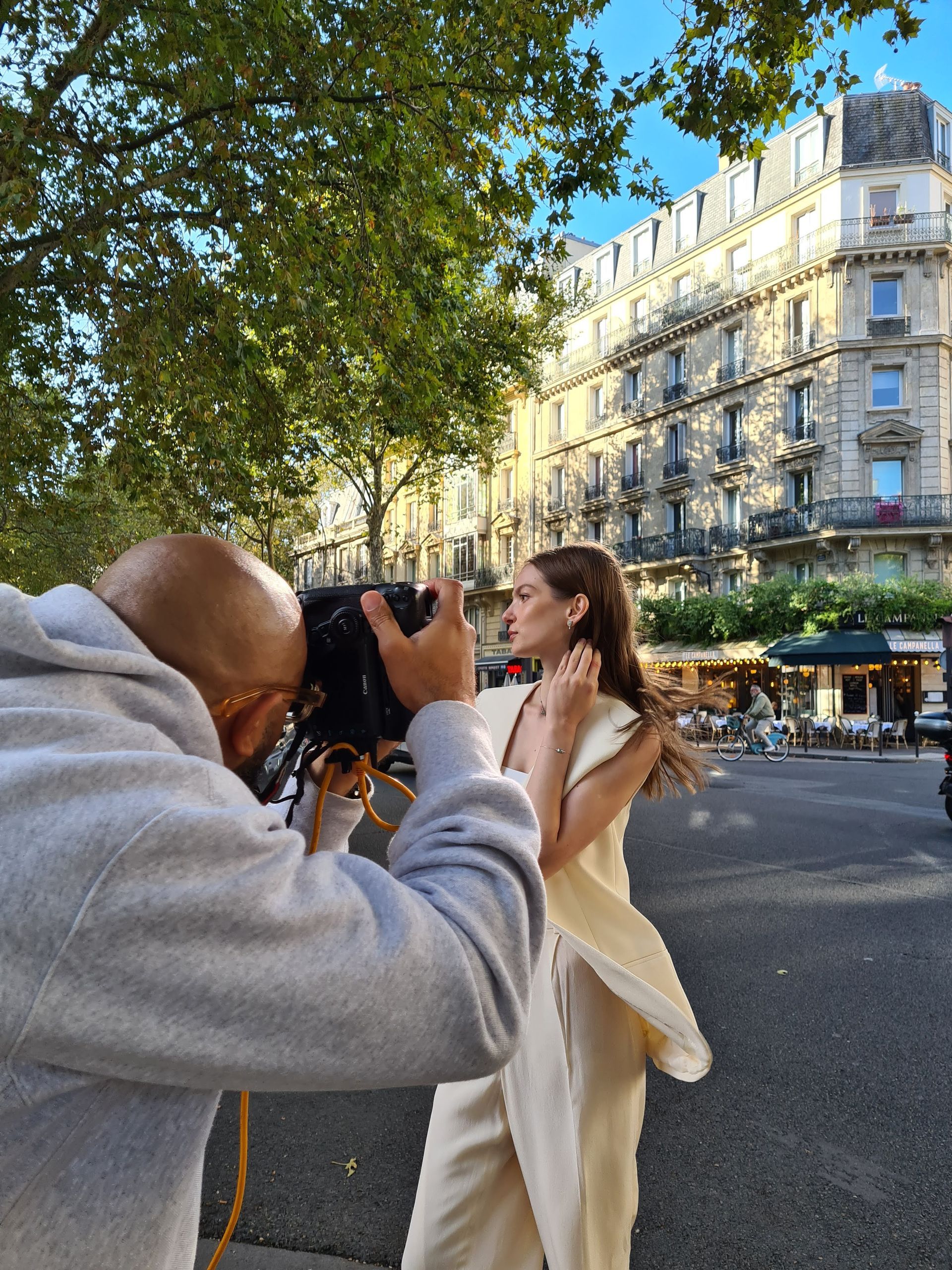 攝影師在城市街道上拍攝一位穿著白色連身裙的人;背景是建築物和樹木。