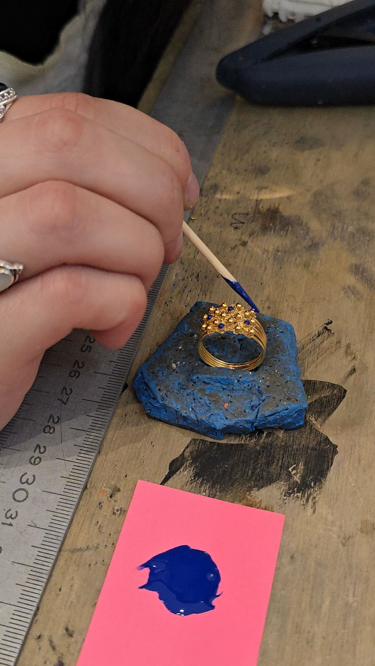一個人用小畫筆沾著粉紅色顏料，在藍色的石頭上把一枚金戒指塗成藍色。