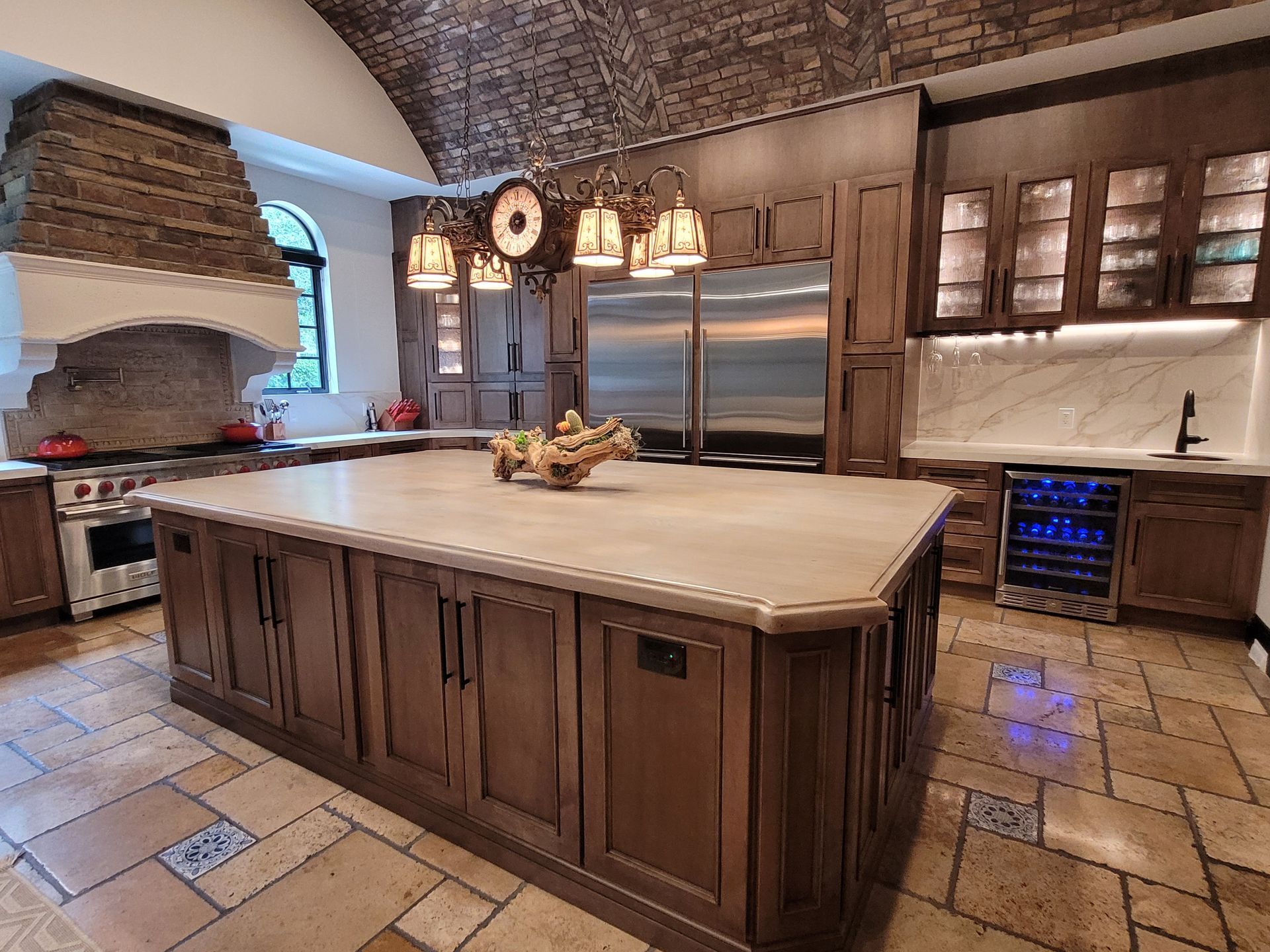 Upscale kitchen with large island, stainless steel fridge, stone range hood, and tile flooring.
