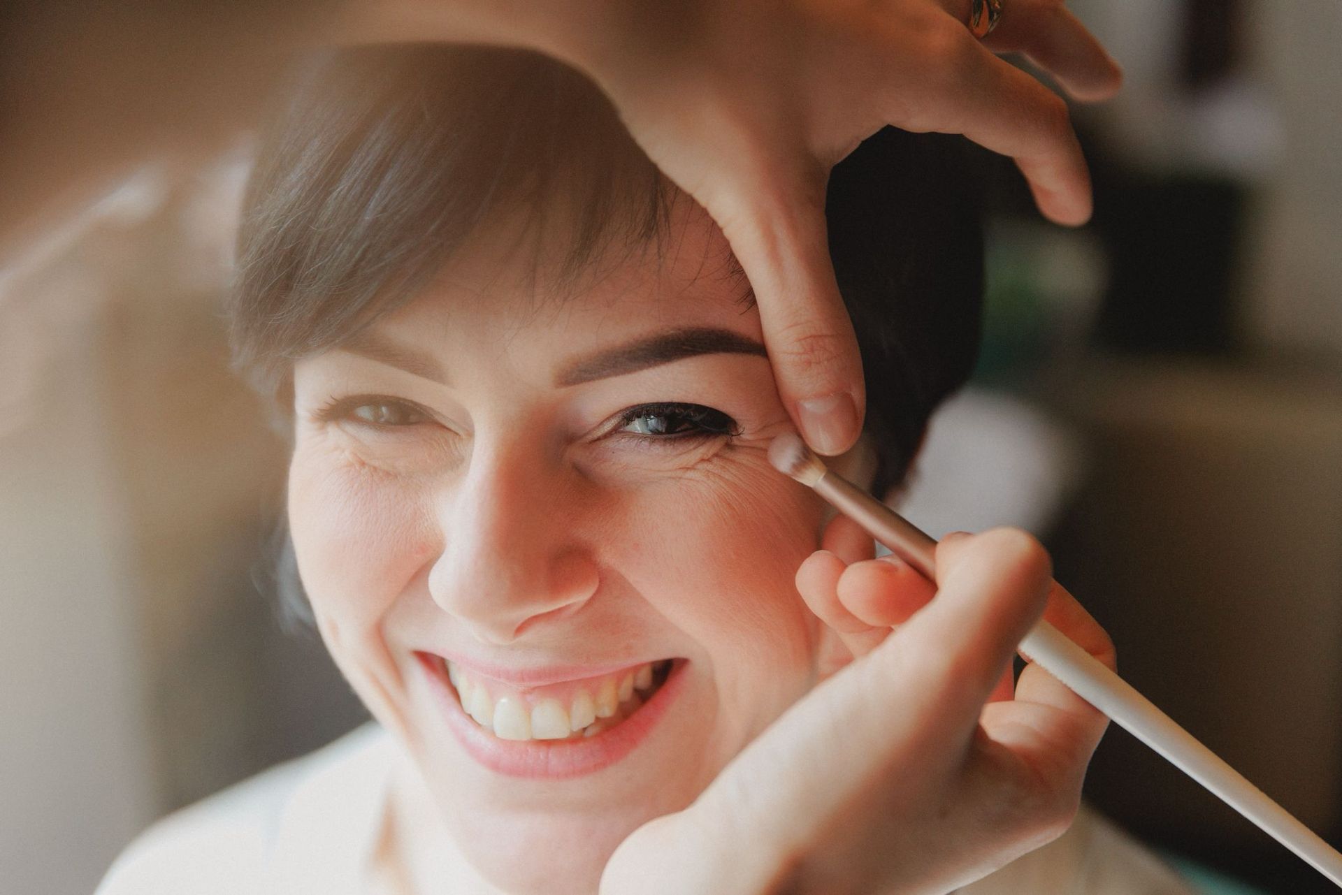 Un truccatore utilizza un pennellino per applicare il correttore vicino all'angolo esterno dell'occhio di una persona sorridente.