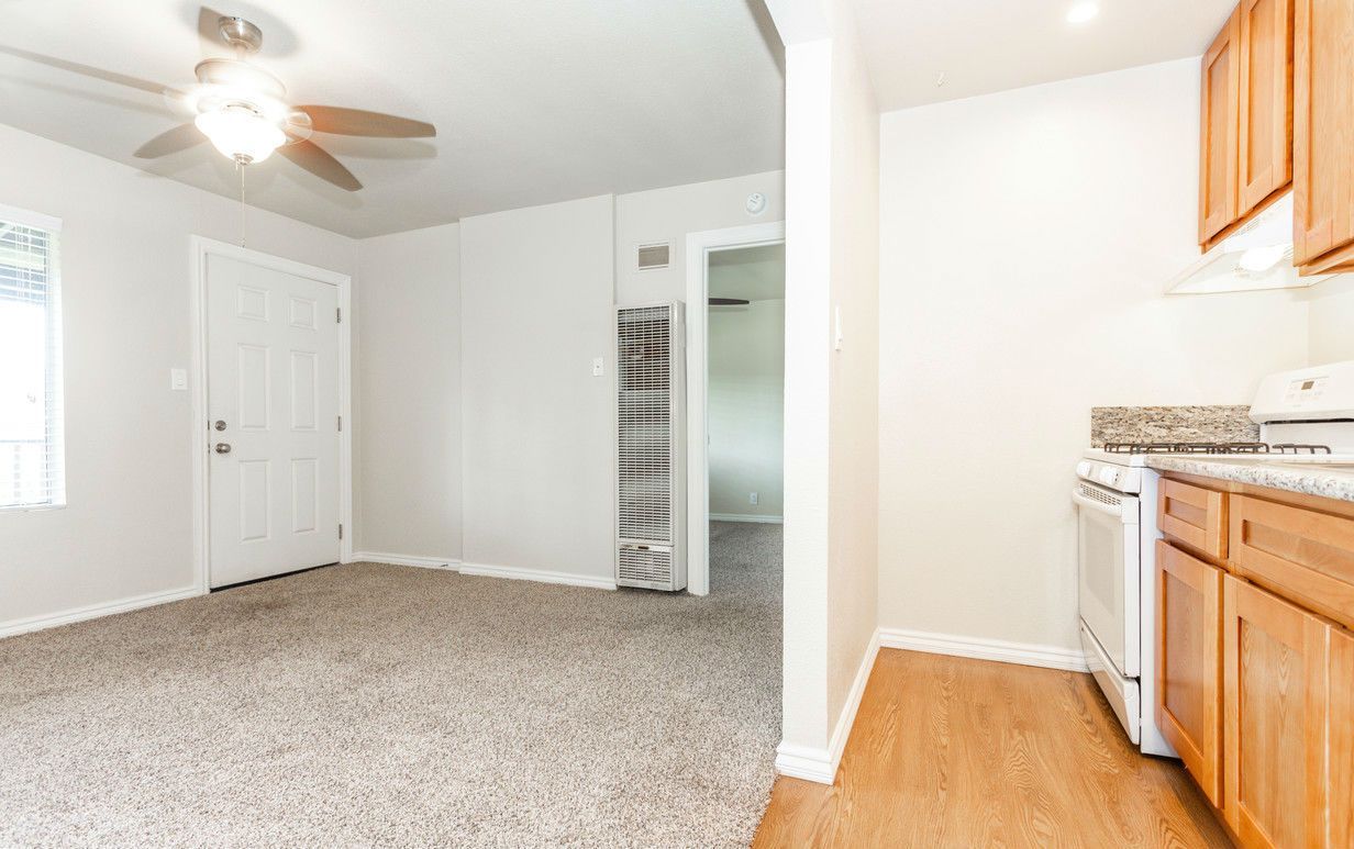 Interior view: living room with tan carpet, white walls, wooden cabinets, and a kitchen.