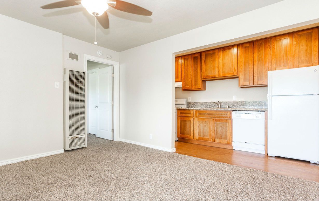 A living room with a kitchen. Light-colored walls, carpet, wooden cabinets, and a white refrigerator.