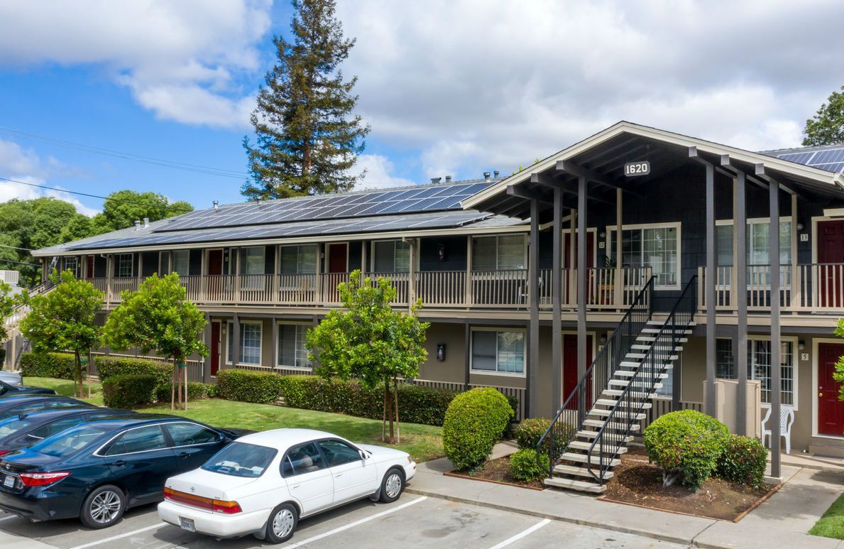Apartment building with cars parked in front, solar panels on the roof, and a stairway. Sunny day.