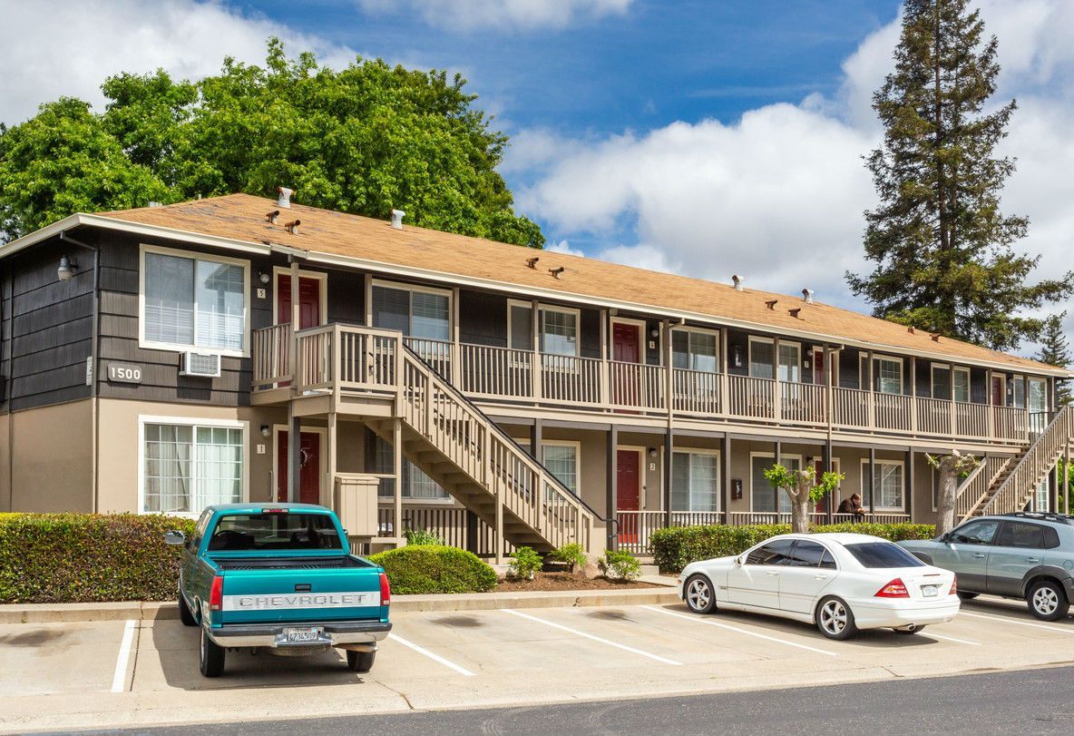 Apartment building with parking, a teal truck, and cars under a cloudy sky.