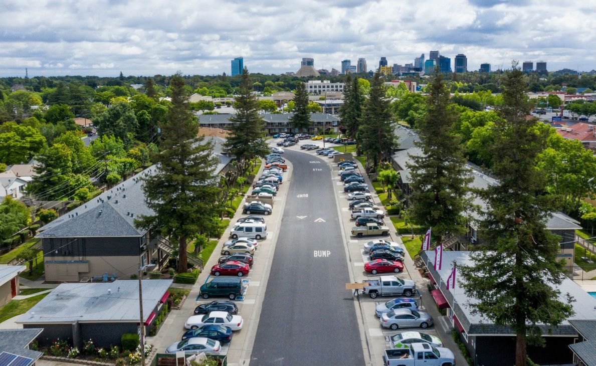Aerial view of a residential street with cars parked along both sides, trees, and a cityscape in the distance.