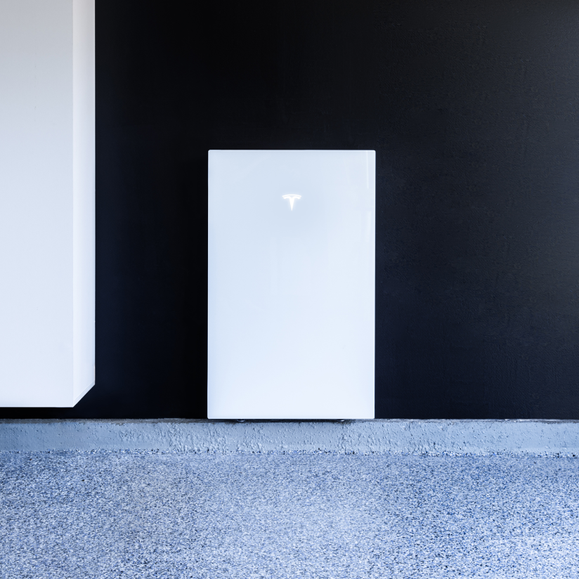 White Tesla Powerwall against black wall, on a speckled gray floor.