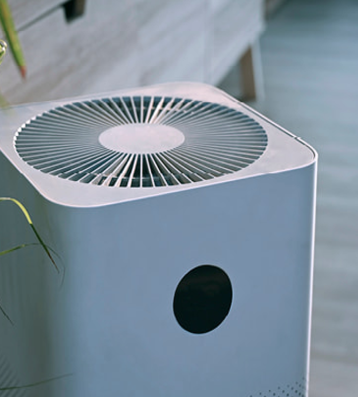 Air purifier, gray, cylindrical with fan top and black circular vent. Indoor setting.