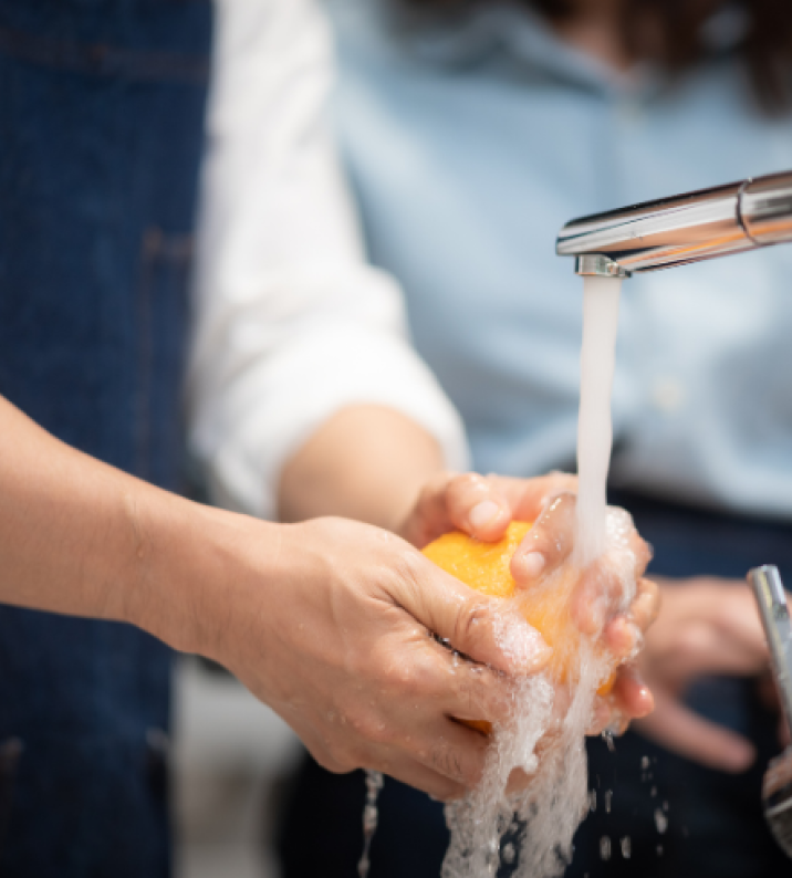 Person washing an orange with soap under running water at a kitchen sink.