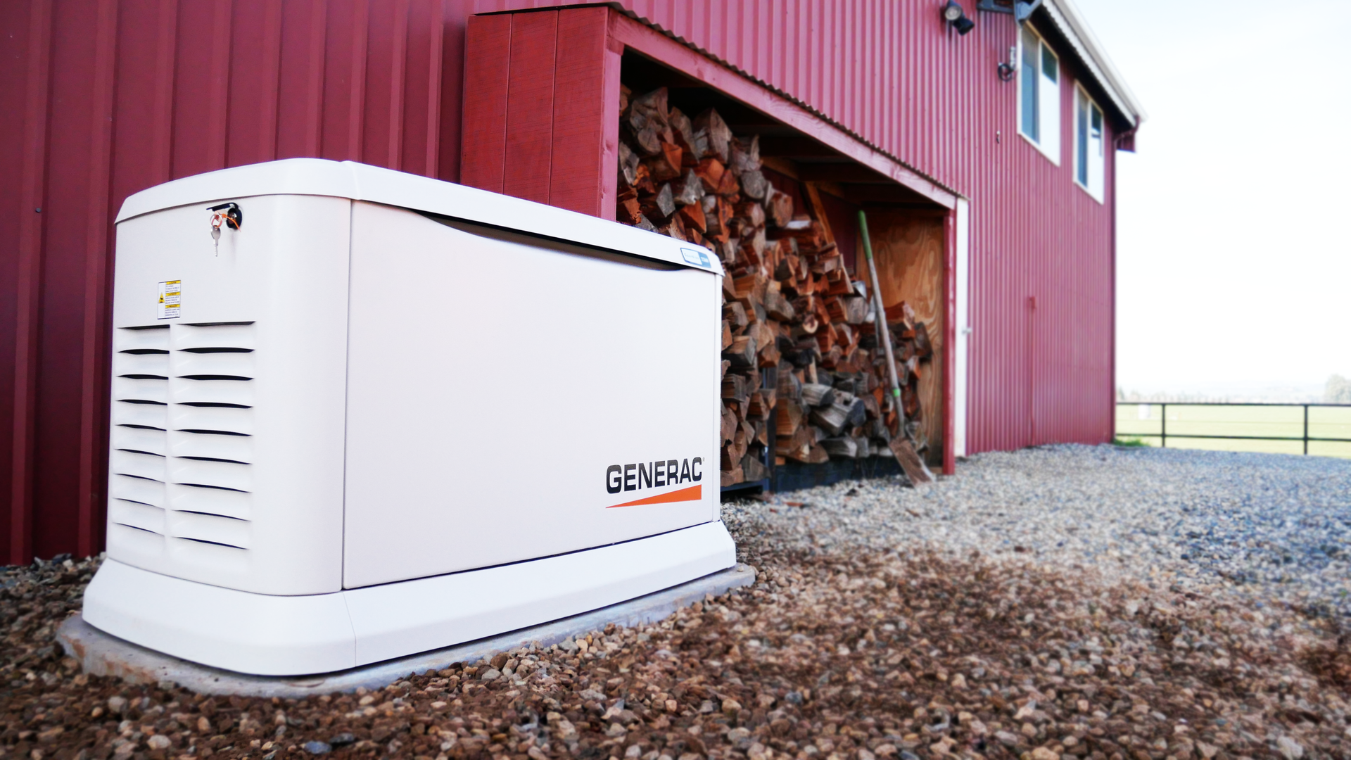 White Generac generator next to a red barn with firewood. Gravel ground.