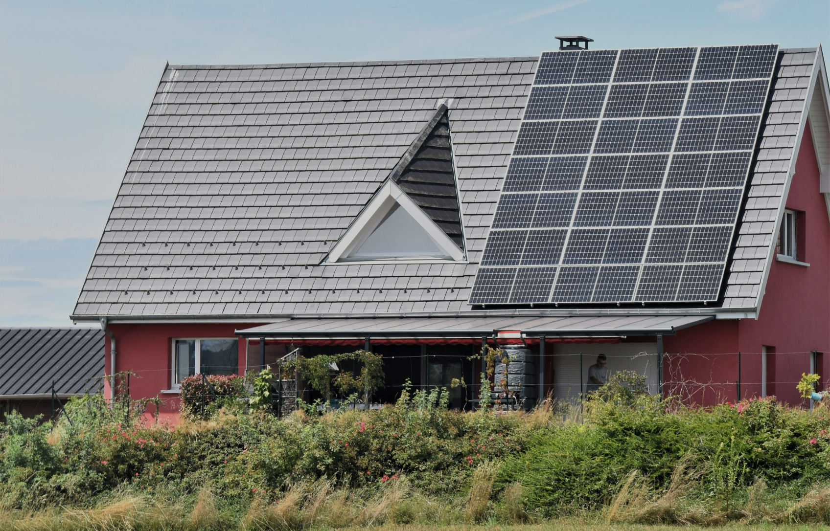 Red house with solar panels on the roof, surrounded by greenery.