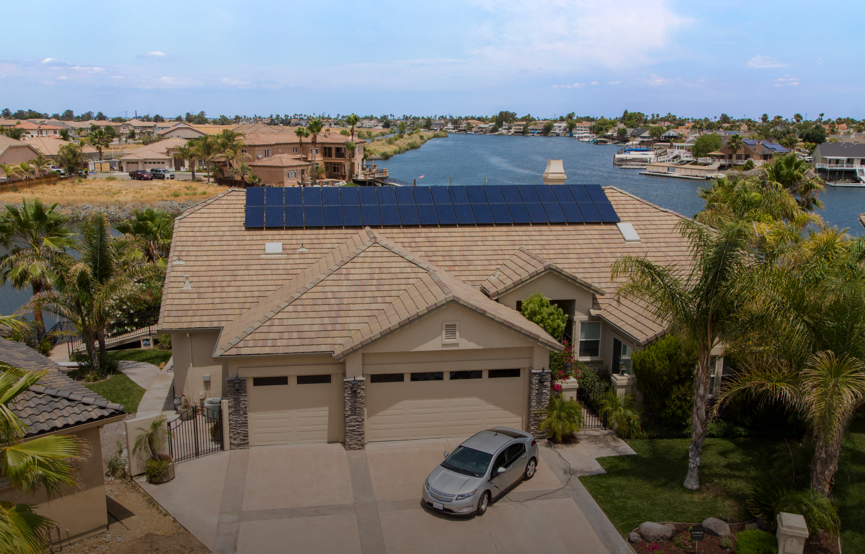 House with solar panels on roof, car in driveway, waterfront setting.