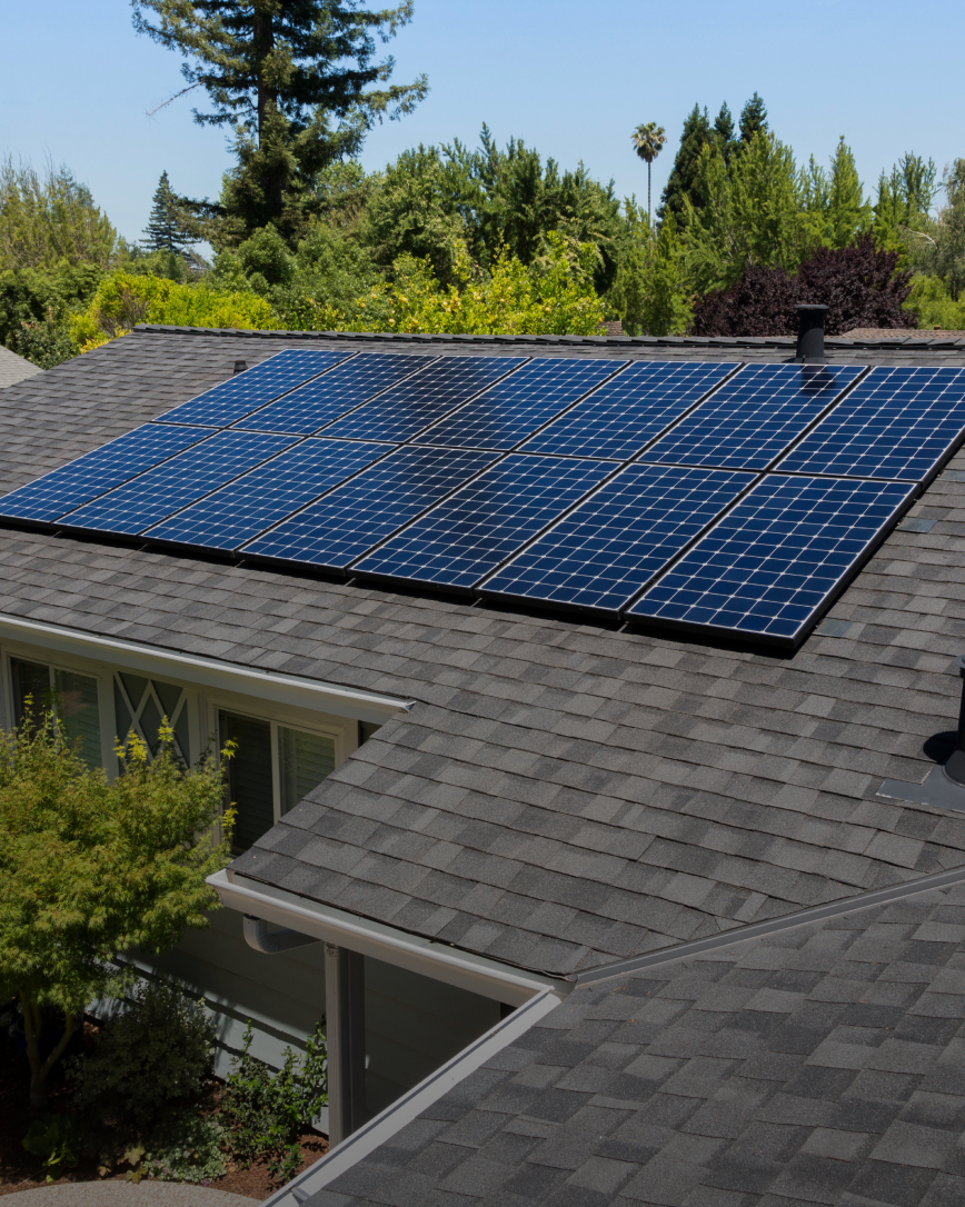 Solar panels installed on a dark gray shingled roof, trees in the background.