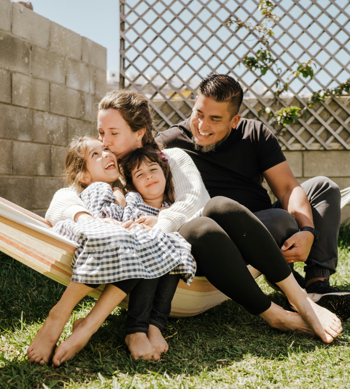 Family of four smiling, relaxing together in a hammock outdoors.