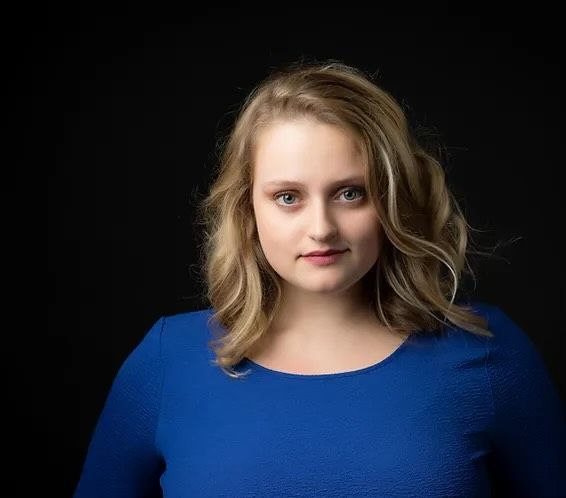 A woman in a blue shirt is standing in front of a black background.