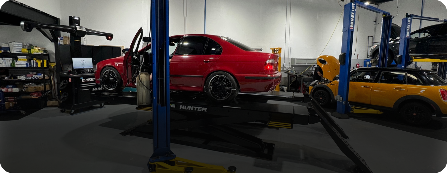 A red car on a lift in a garage, next to a yellow SUV on a lift. Blue lift columns and tools visible.