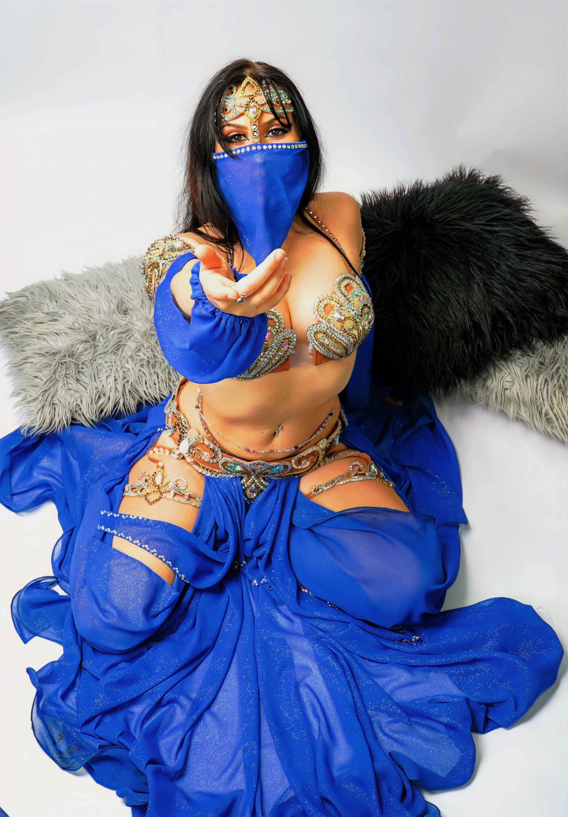 Belly dancer Azeeria Azizah in a blue dress and mask is sitting on a bed.