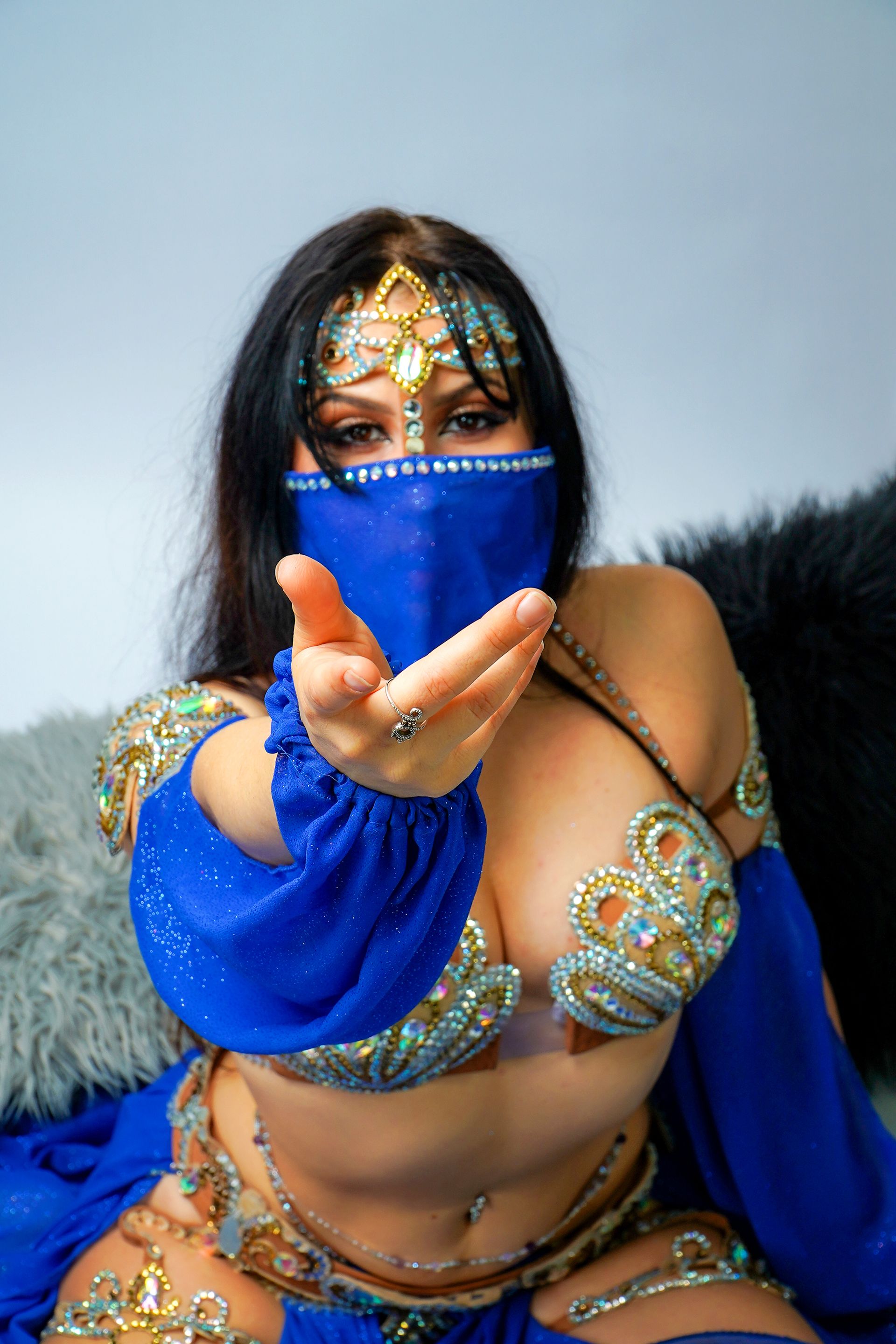Belly dancer Azeeria Azizah in a blue and gold belly dancer costume is sitting on a couch.