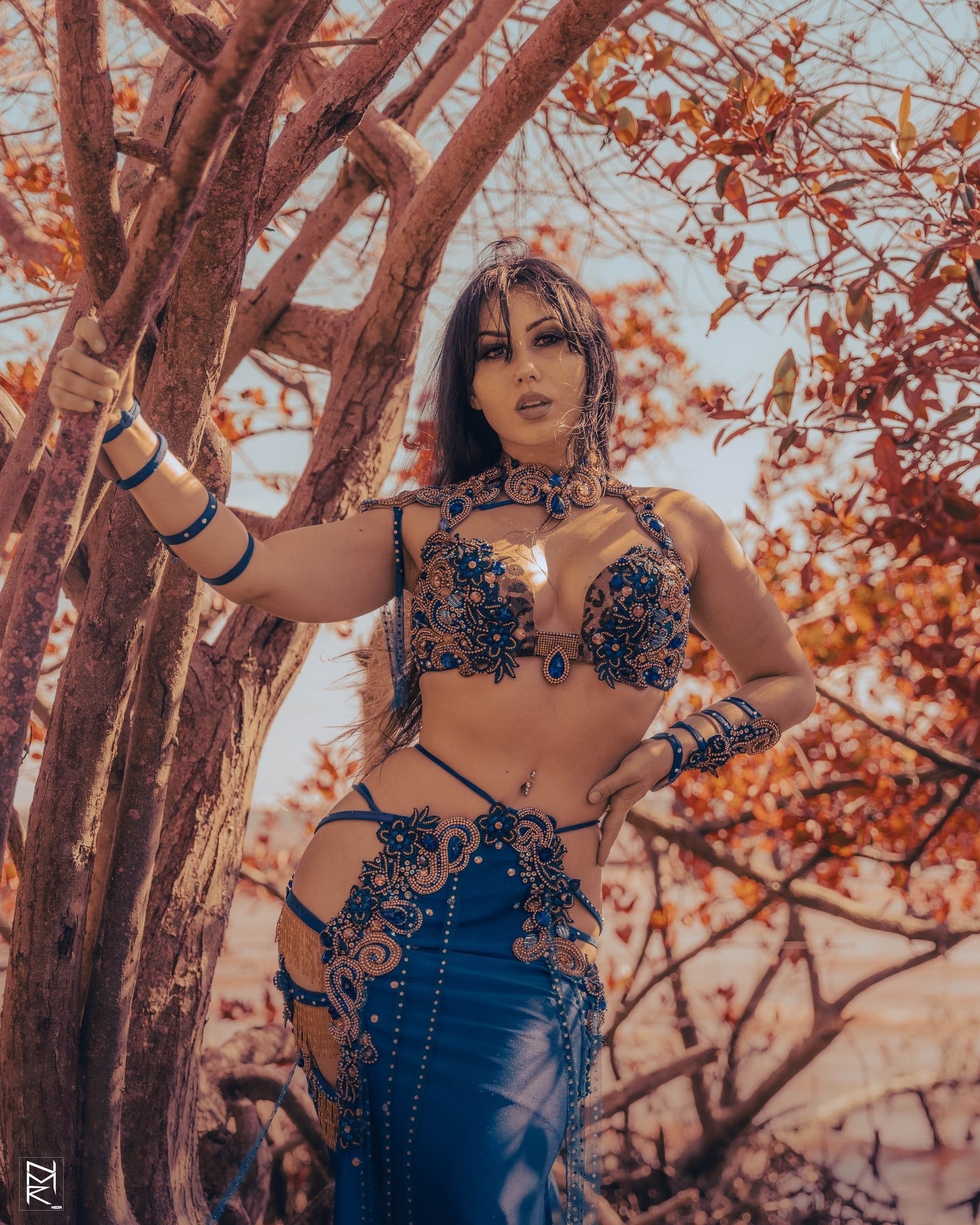 Belly dancer Azeeria Azizah in a blue dress is standing next to a tree.
