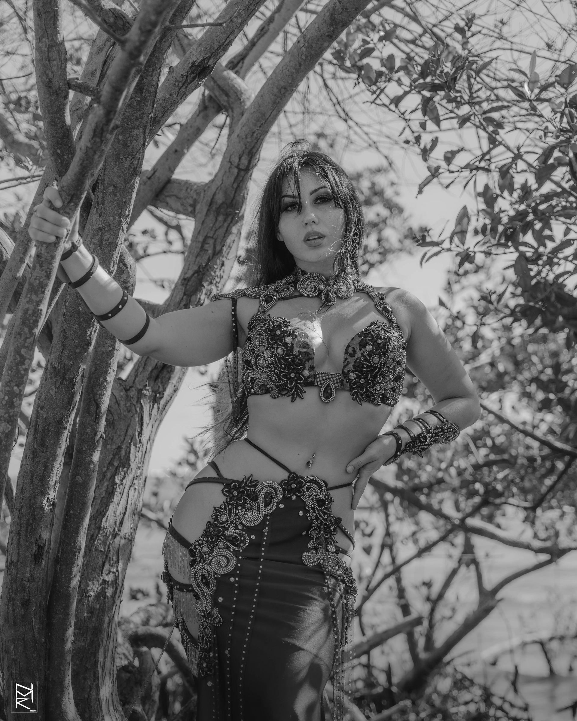 A black and white photo of Belly dancer Azeeria Azizah in a belly dancer costume standing next to a tree.