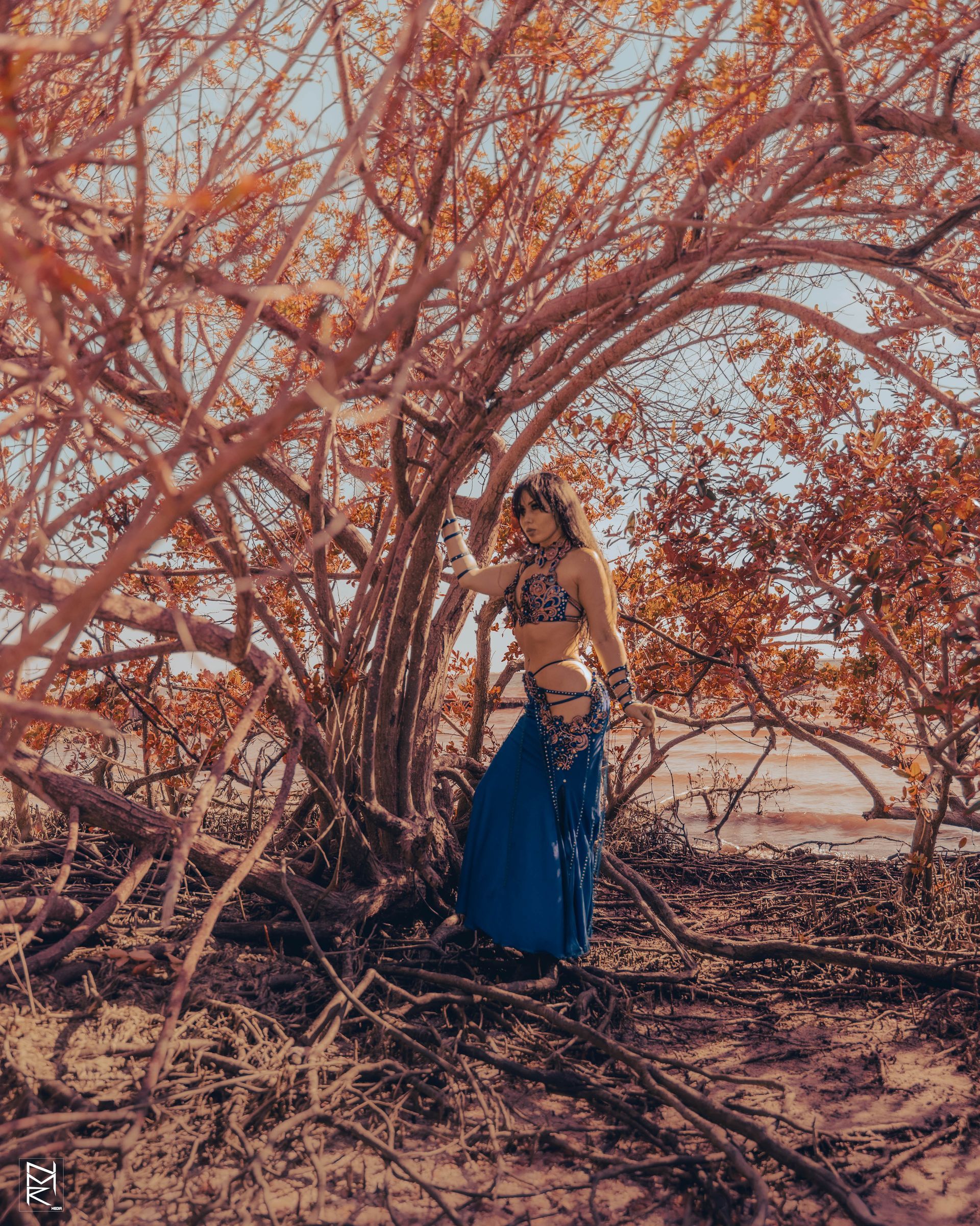 Azeeria Azizah in a blue dress is standing under a tree posing with one hand holding a branch.