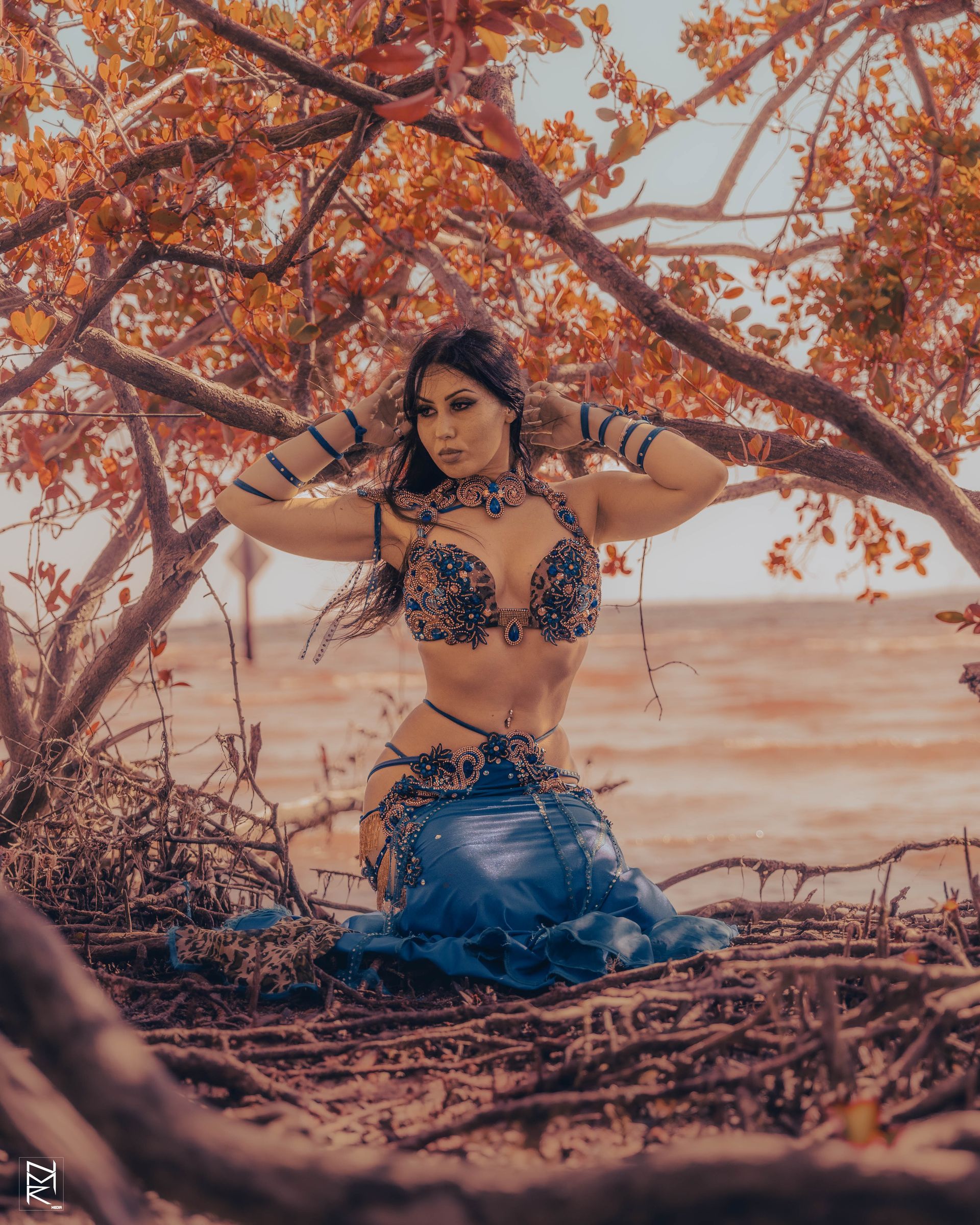 Belly dancer Azeeria Azizah in a belly dance costume is kneeling under a tree.
