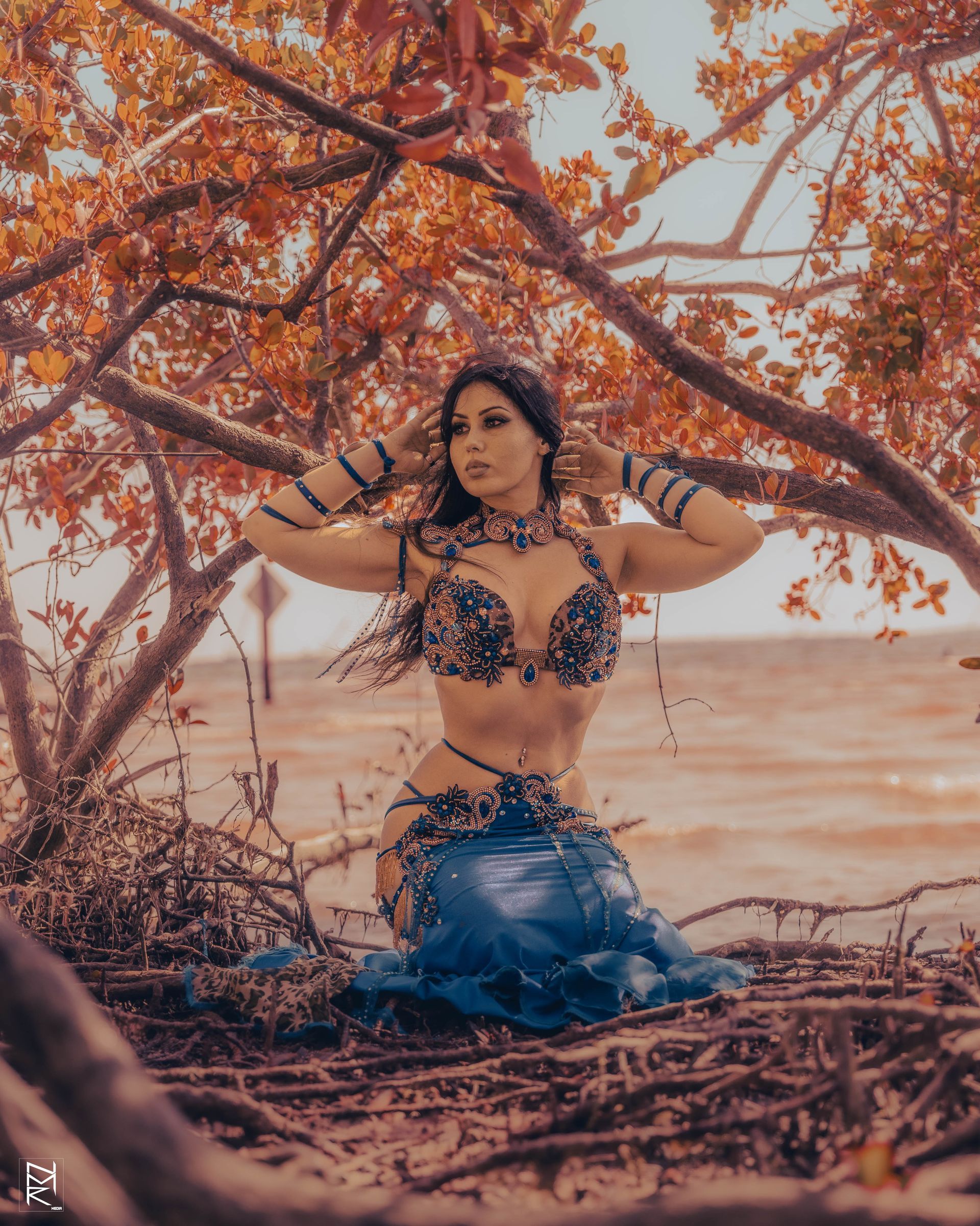 Belly dancer Azeeria Azizah is kneeling under a tree on the beach.