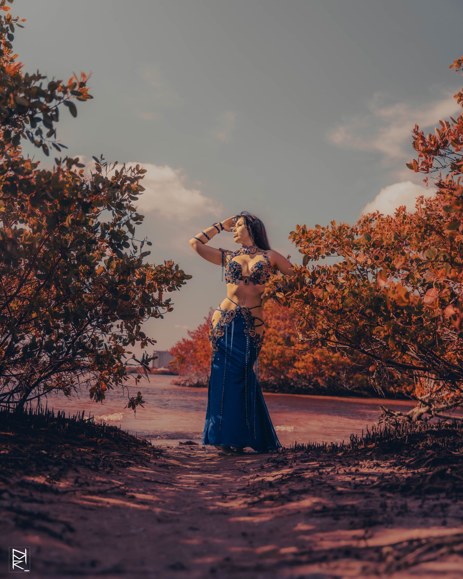 Azeeria Azizah in a blue dress is standing in the middle of the sand between trees.