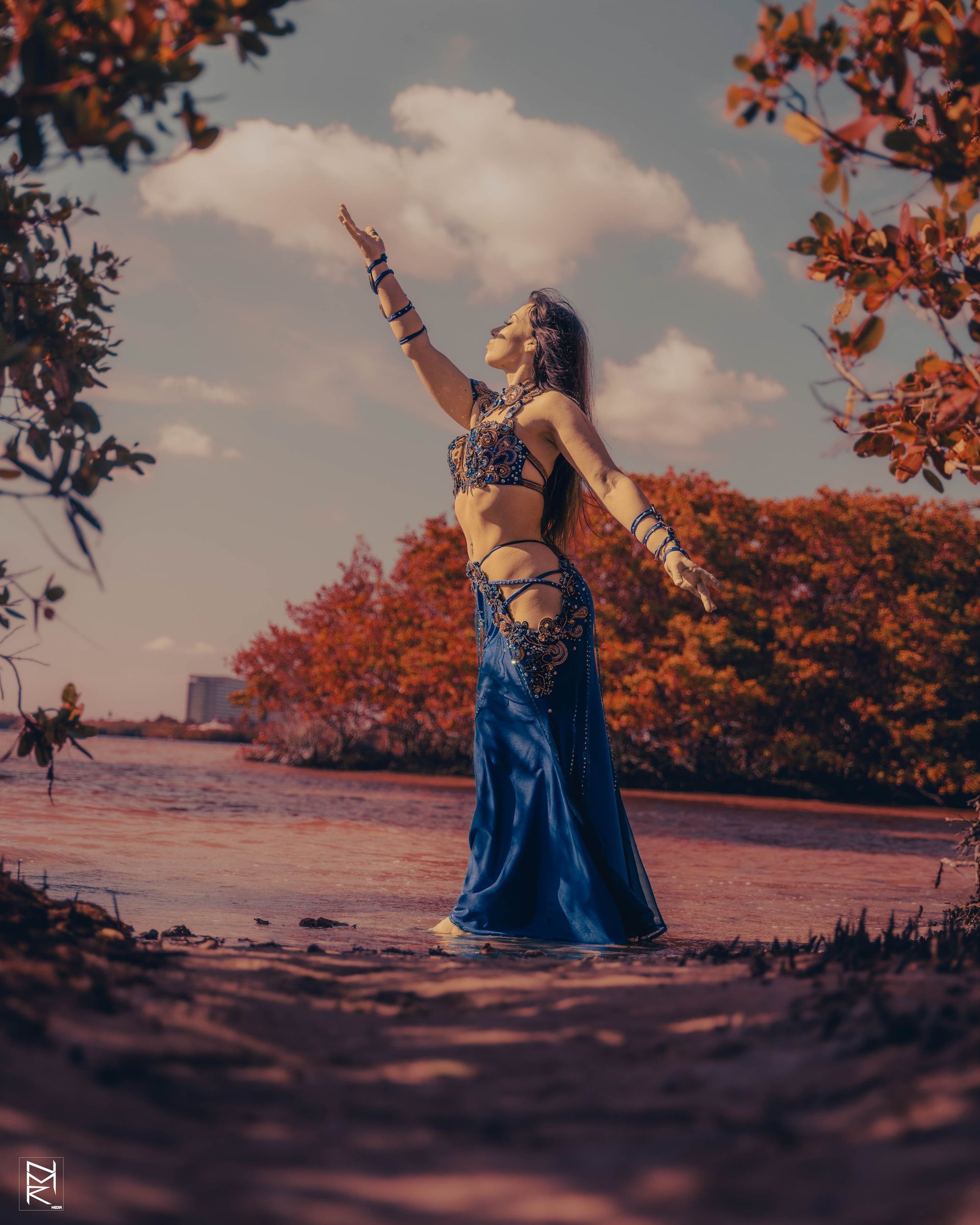 Belly Dancer Azeeria Azizah in a blue dress is standing in front of a body of water with her arms outstretched.