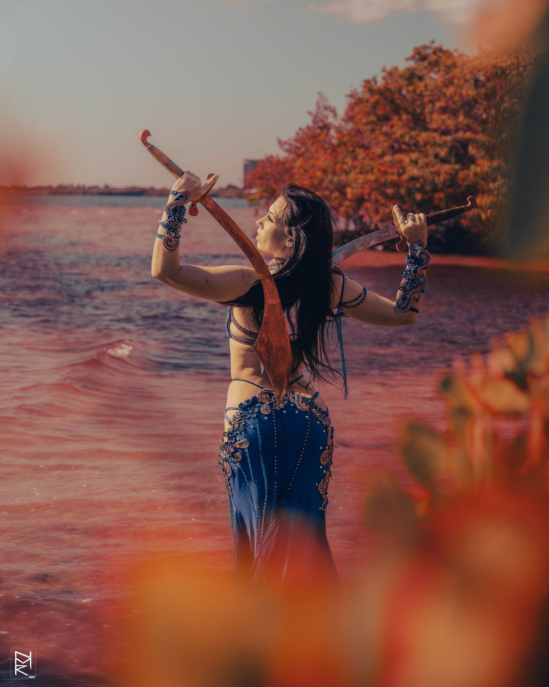 Bellydancer Azeeria Azizah standing in water holding two swords over her back.