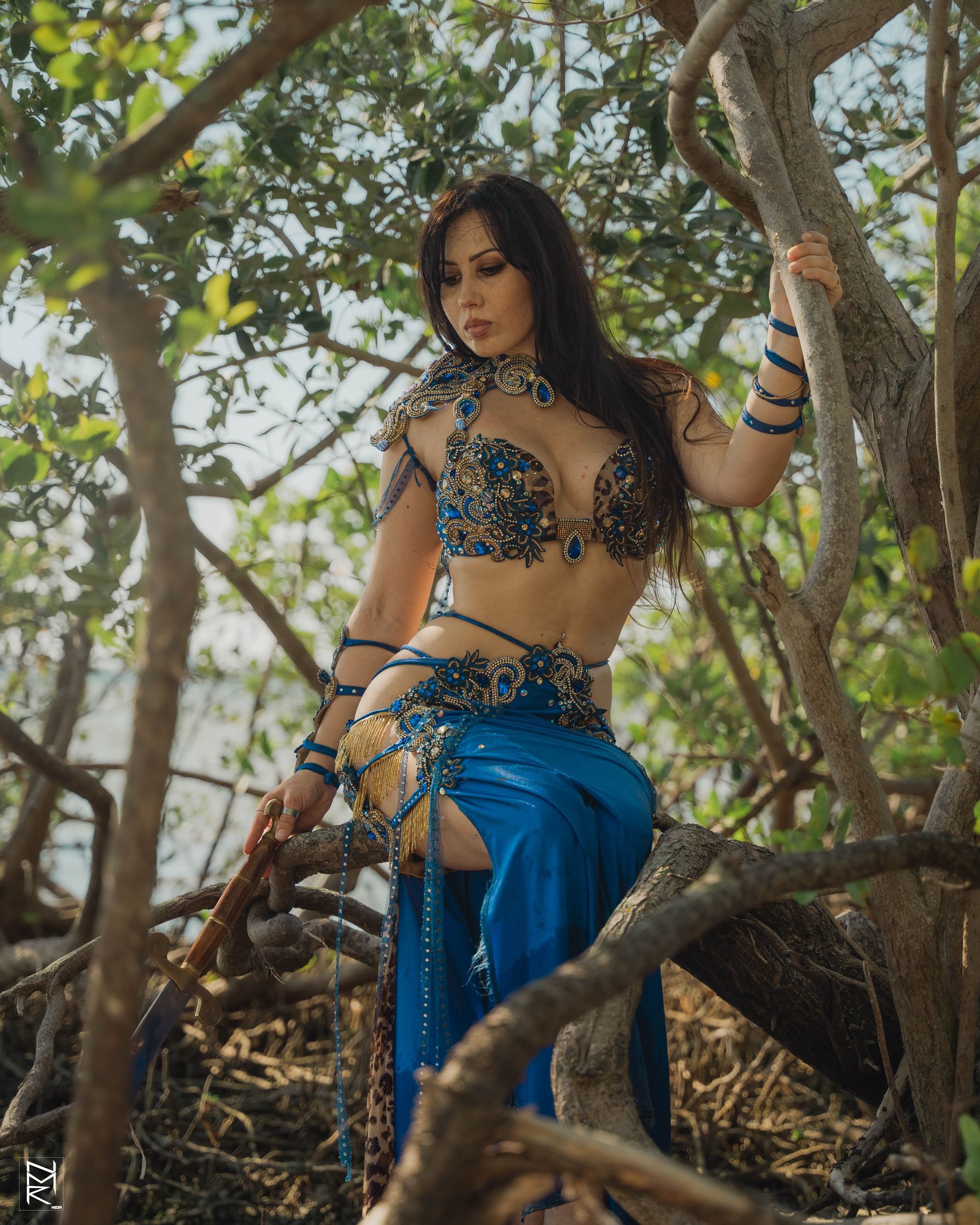Belly dancer Azeeria Azizah in a belly dancer costume is sitting on a tree branch.