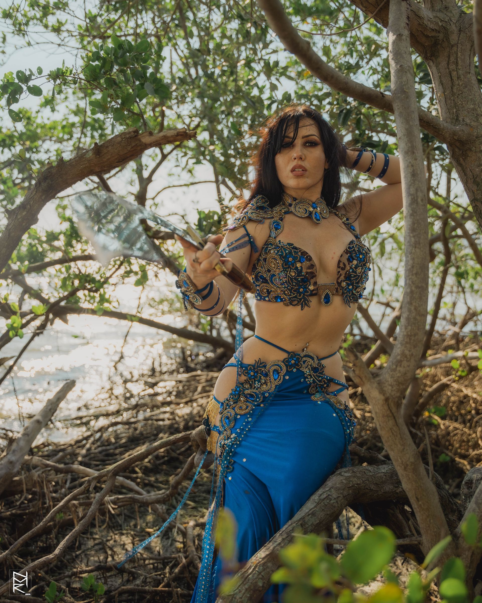 Belly dancer Azeeria Azizah in a blue dress is sitting on a tree branch with her hand out.
