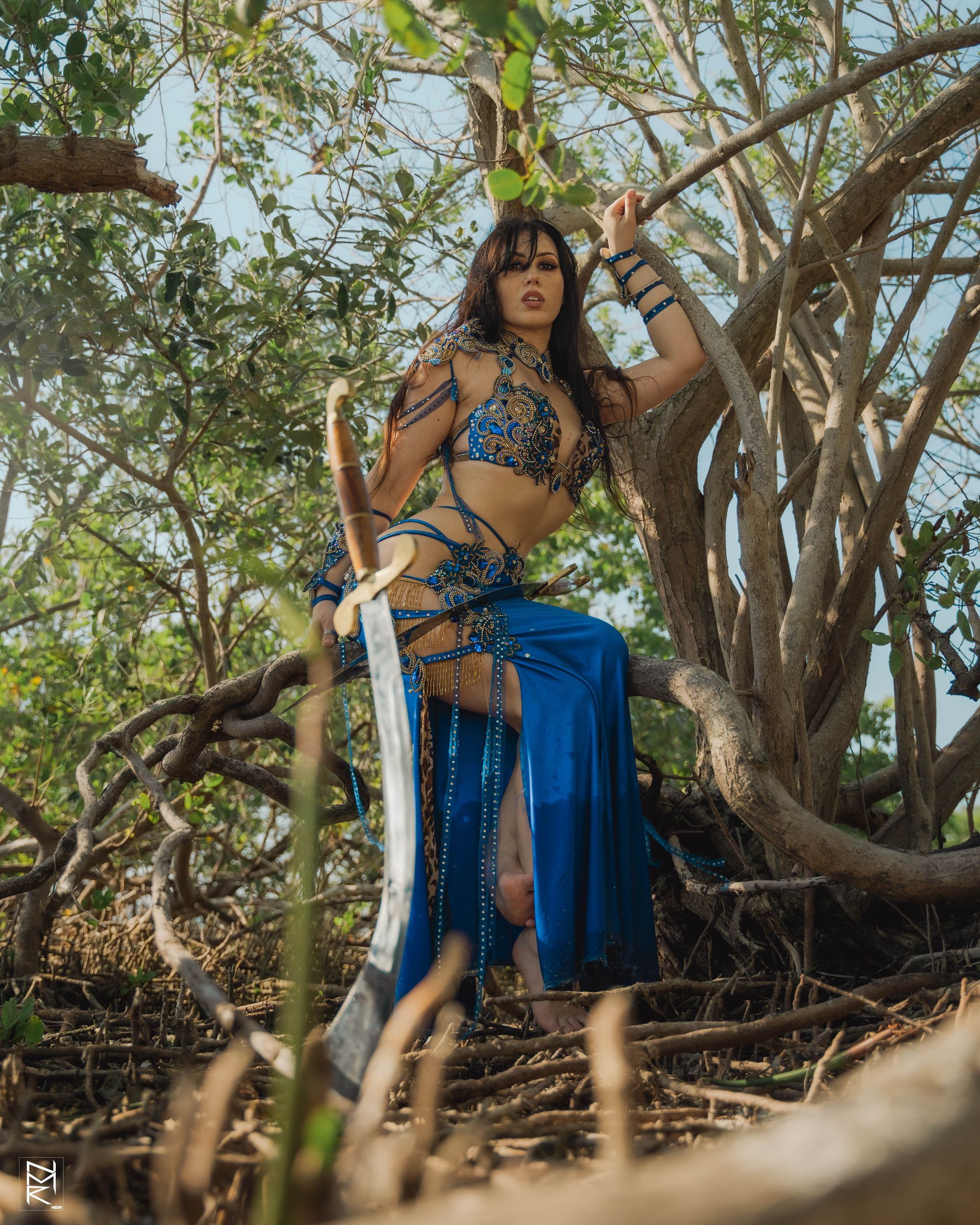 Belly dancer Azeeria Azizah in a blue dress is sitting on a tree branch.