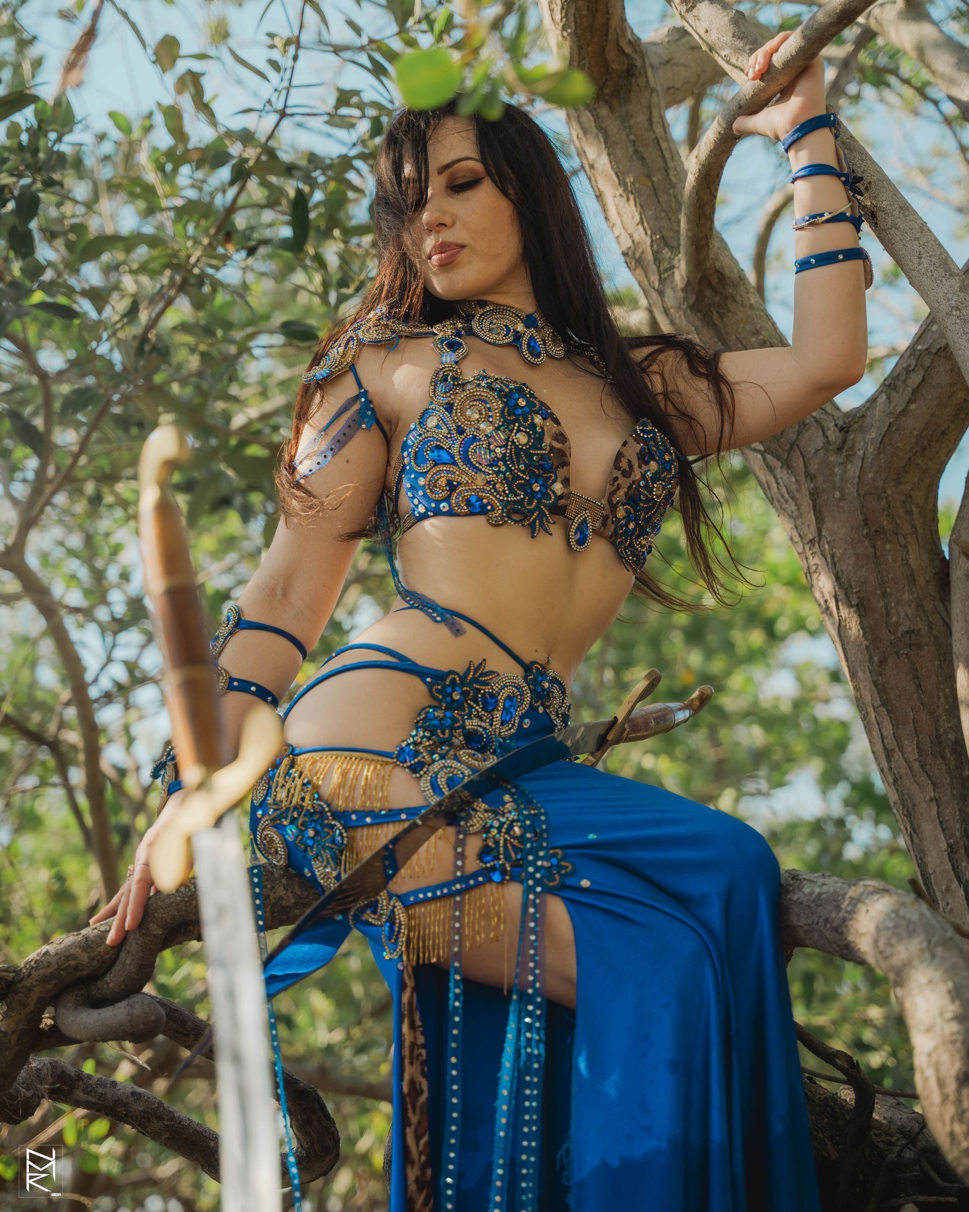 Belly dancer Azeeria Azizah in a blue dress is sitting on a tree branch.