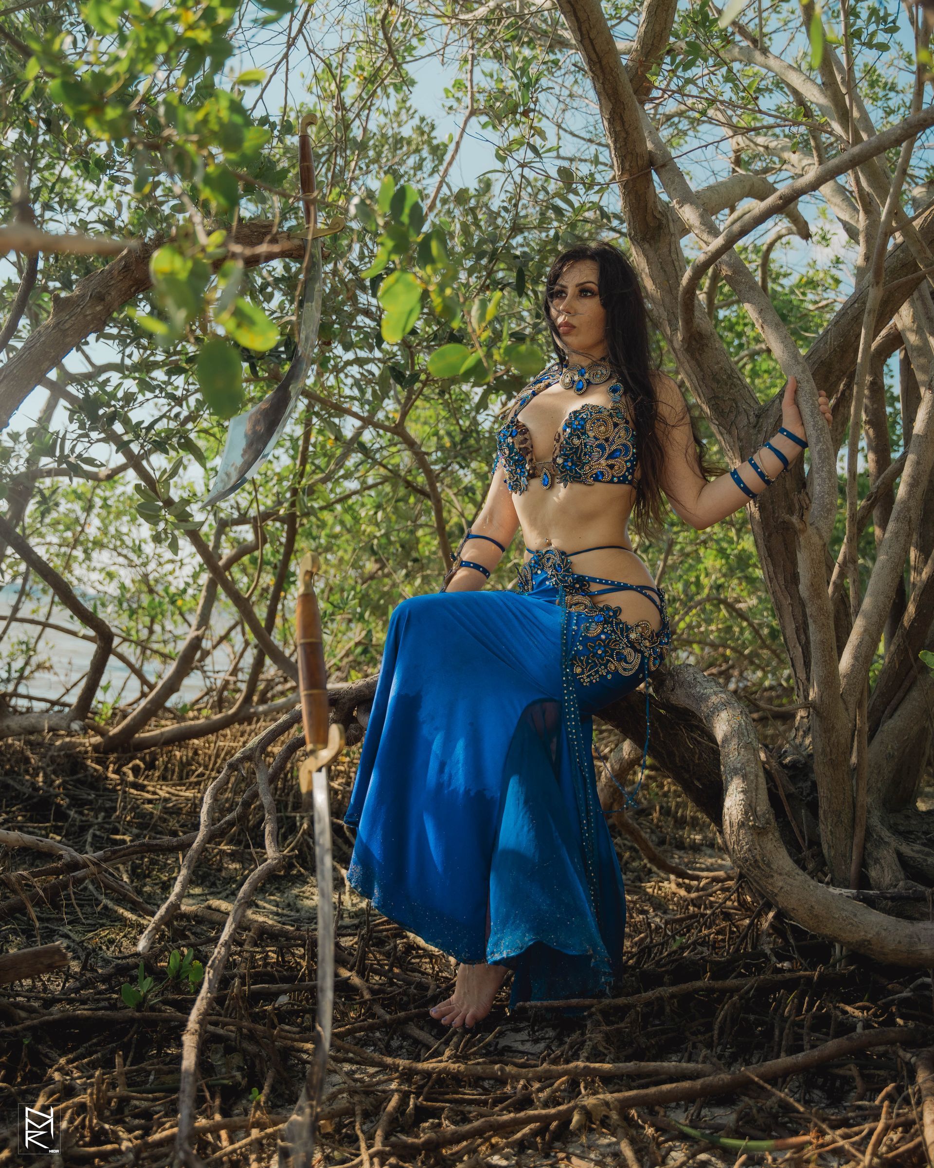 Belly dancer Azeeria Azizah in a blue dress is sitting on a tree branch.